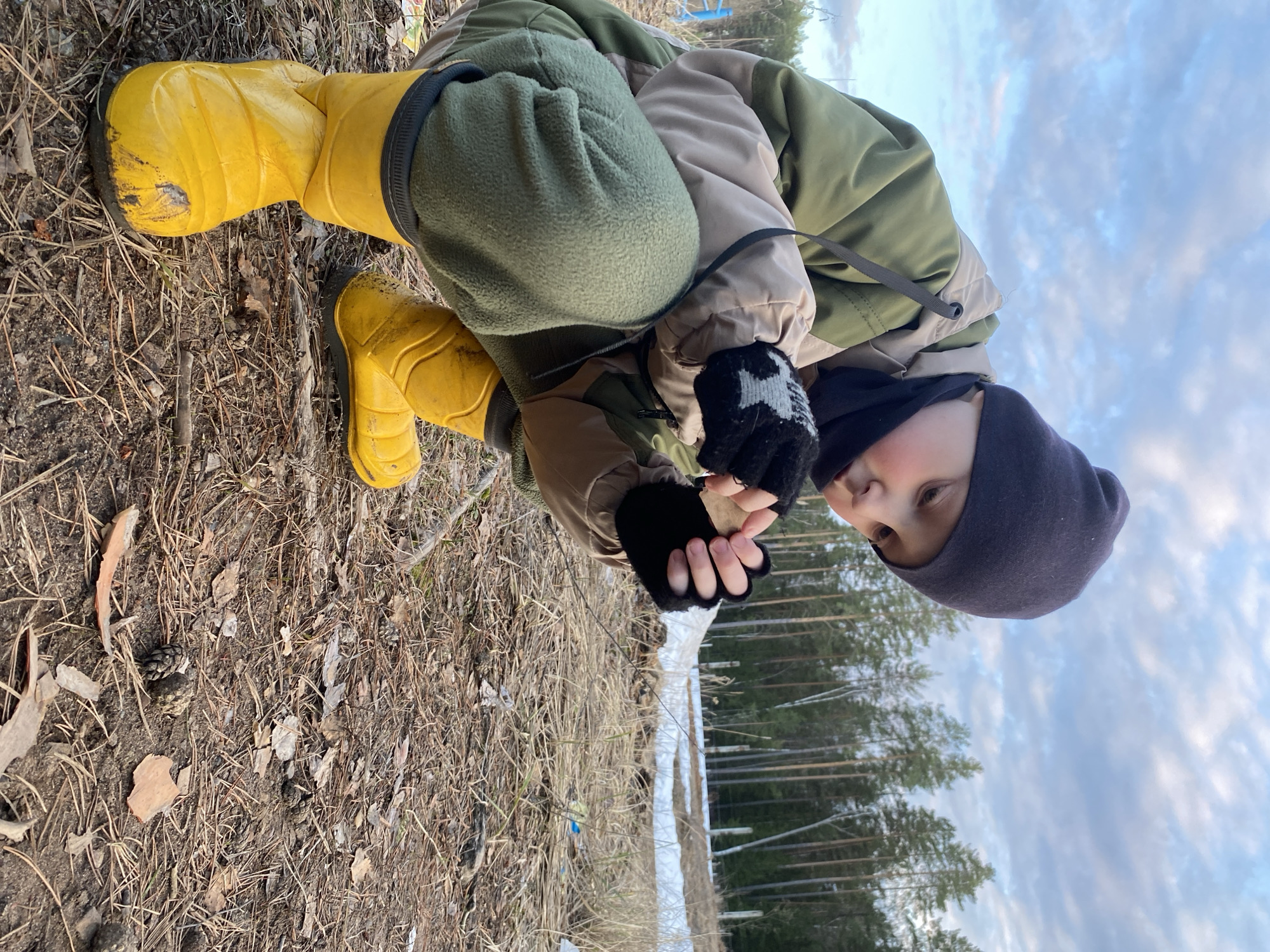 A young child wearing yellow rubber boots, olive green trousers, and a multi-toned beige and green jacket with black gloves and a black beanie is crouching near a patch of ground covered in dried leaves and twigs. The child is holding a small, undefined object in their gloved hands. In the background is a field with patches of snow and a distant treeline under a partly cloudy sky. The location is identified as being near Noviy, Russia.