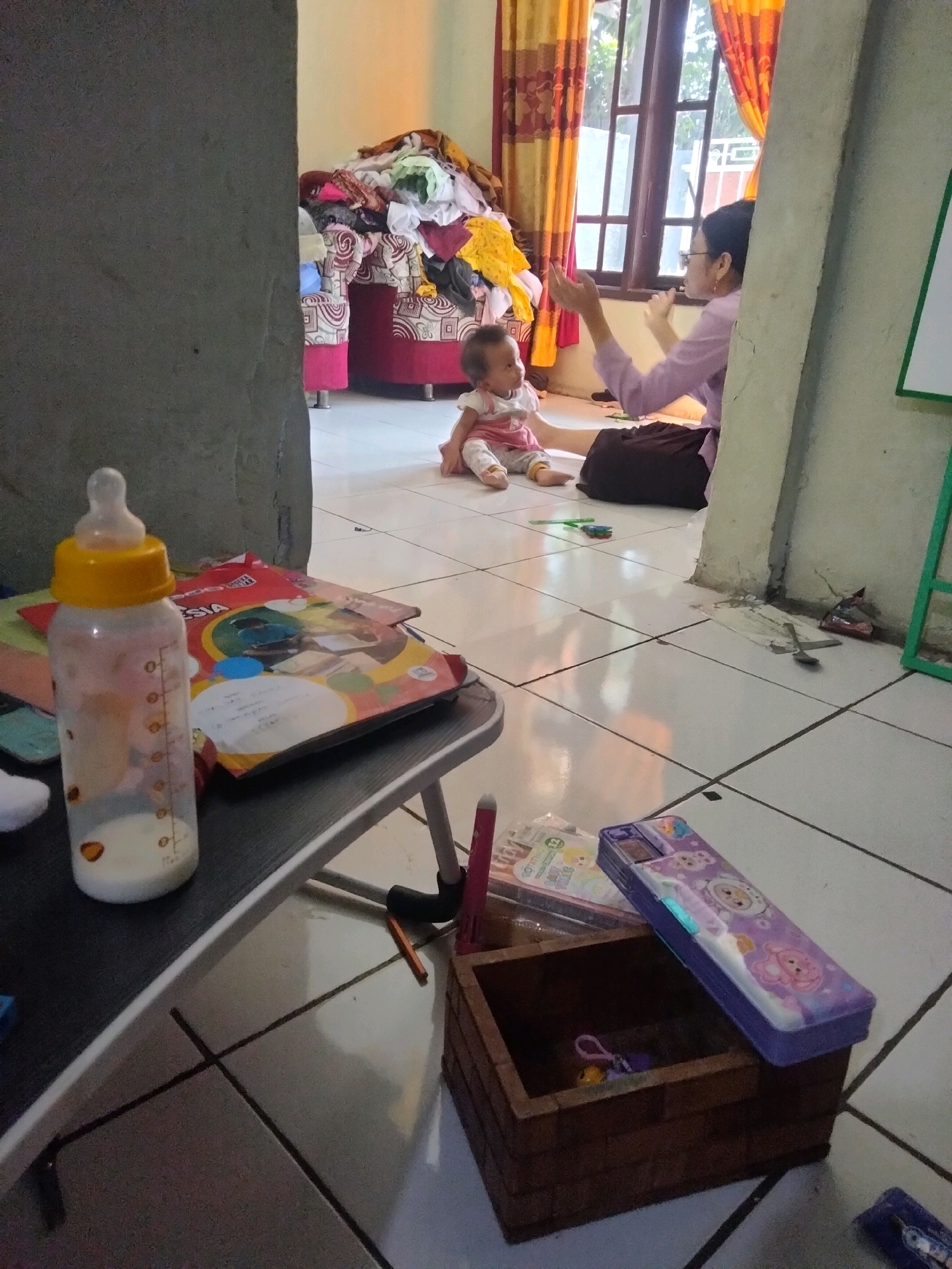 In the city of Suka Sari, Indonesia, a dimly lit indoor scene unfolds. A young child, dressed in a pink and white outfit, sits on a tiled floor in the foreground. To the child's right, an adult wearing glasses and a lavender shirt is seated, their hands raised in what appears to be a playful interaction with the child. Behind them, a window is adorned with orange and yellow curtains, and a pile of colorful clothing is visible on a red sofa.
To the left of the child, a baby bottle filled with milk stands on a small table, next to a stack of books. In the foreground on the right, a wooden box holds a small purple object. A purple pencil case with cartoon characters rests on top of the box. The floor is a white tiled surface, with dark grout lines. The overall impression is of a domestic environment, likely a living room or playroom.