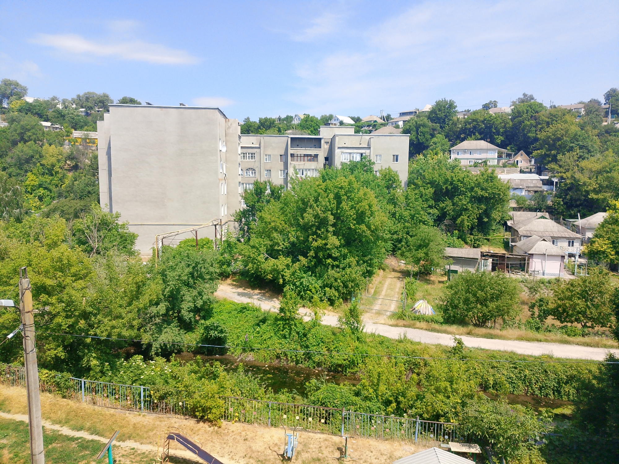 The image depicts an overcast sky with scattered clouds above a residential area in Rîbniţa, Moldova. In the foreground, a metal fence runs horizontally, bordering a dry, grassy area with a children's slide visible. Beyond the fence, a narrow body of water, possibly a stream or canal, is flanked by dense green foliage. In the mid-ground, a concrete utility pole with wires is positioned on the left side. Further back, a multi-story apartment building with a plain grey facade dominates the left half of the frame, partially obscured by lush green trees. To the right of the apartment building, a series of smaller residential houses with pitched roofs are scattered across a sloping, tree-covered hillside. A dirt road or path winds through the landscape. The overall scene suggests a mixed urban and natural environment, characterized by residential structures and abundant vegetation under a bright sky.