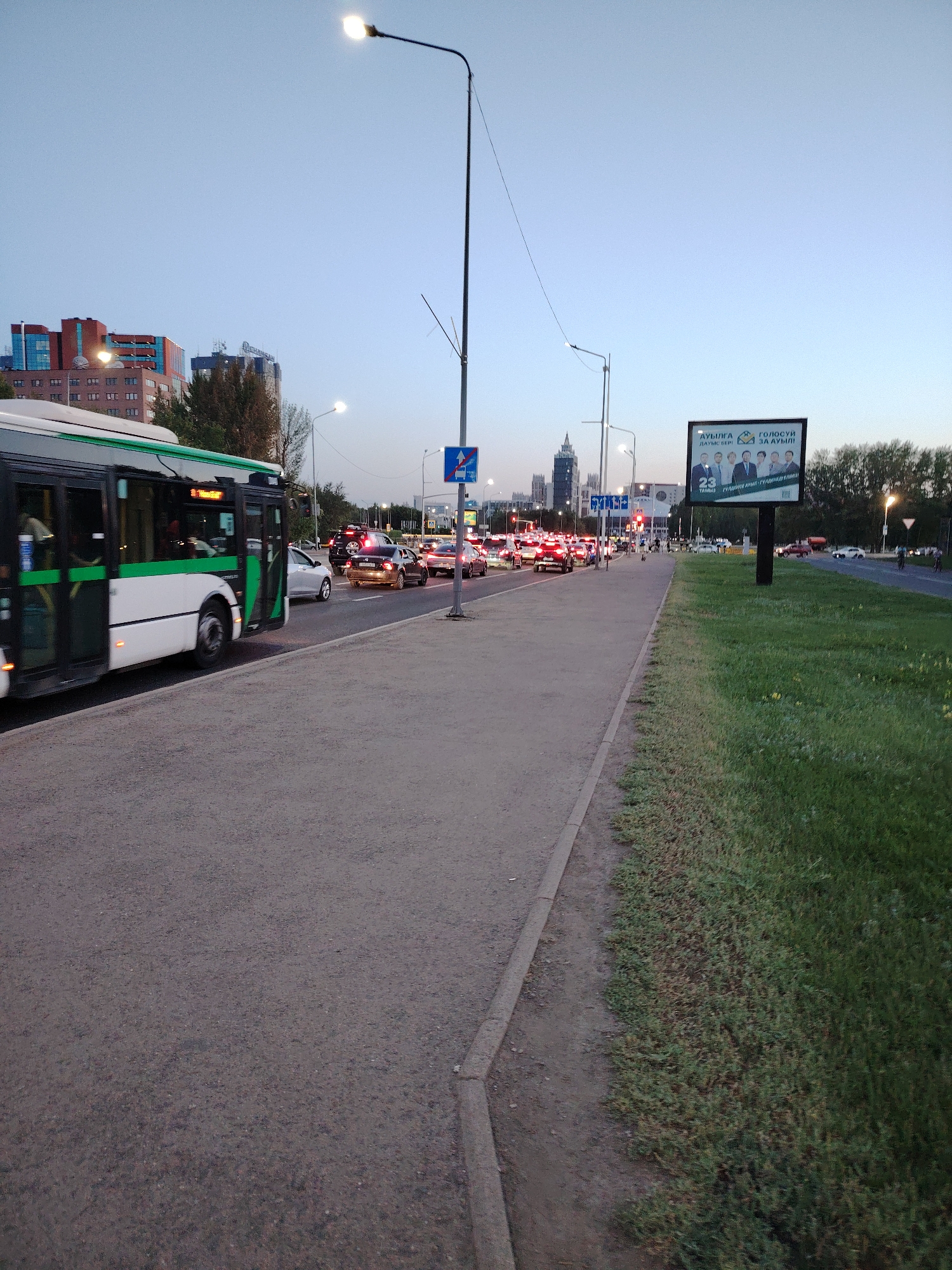 The image depicts a street scene in Astana, Kazakhstan, during twilight. A white and green bus is moving on the left side of the frame, while a line of cars is visible in traffic on the right. There is a pedestrian walkway alongside the road, with a grassy area and trees beyond. A large billboard with an election advertisement featuring several men in suits is prominently displayed on the right. Streetlights are illuminated, and the sky is a clear blue. The overall atmosphere is calm, despite the presence of traffic.