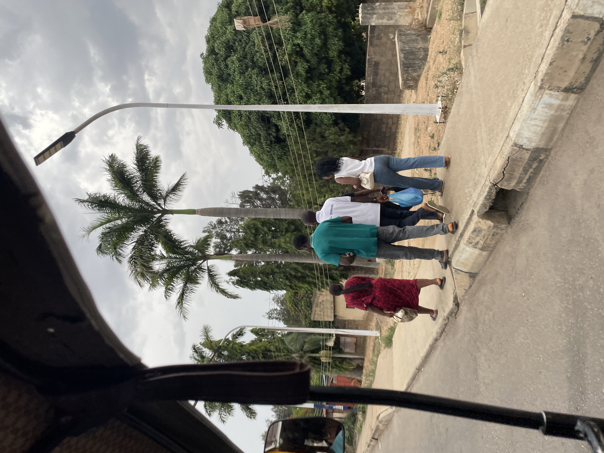 The image depicts a street scene in Jos, Nigeria, observed from inside a vehicle, possibly a car or auto-rickshaw, given the view through the front window and frame. Four people are walking along a paved sidewalk on the right side of the frame. Starting from the furthest person, a woman in jeans and a white shirt is holding a white bag. Behind her, a person wears a white shirt with blue text, possibly part of a uniform or branded clothing. A man in a green shirt is walking next in line. The closest person in the procession is a woman in a red patterned dress carrying a light-colored bag.
A palm tree is prominent in the background, along with other trees and utility poles. The sky is overcast with gray clouds. The sidewalk is bordered by a concrete curb. In the lower portion of the frame, the vehicle's dashboard, rearview mirror, and windshield frame are visible, indicating the perspective is from inside a vehicle.