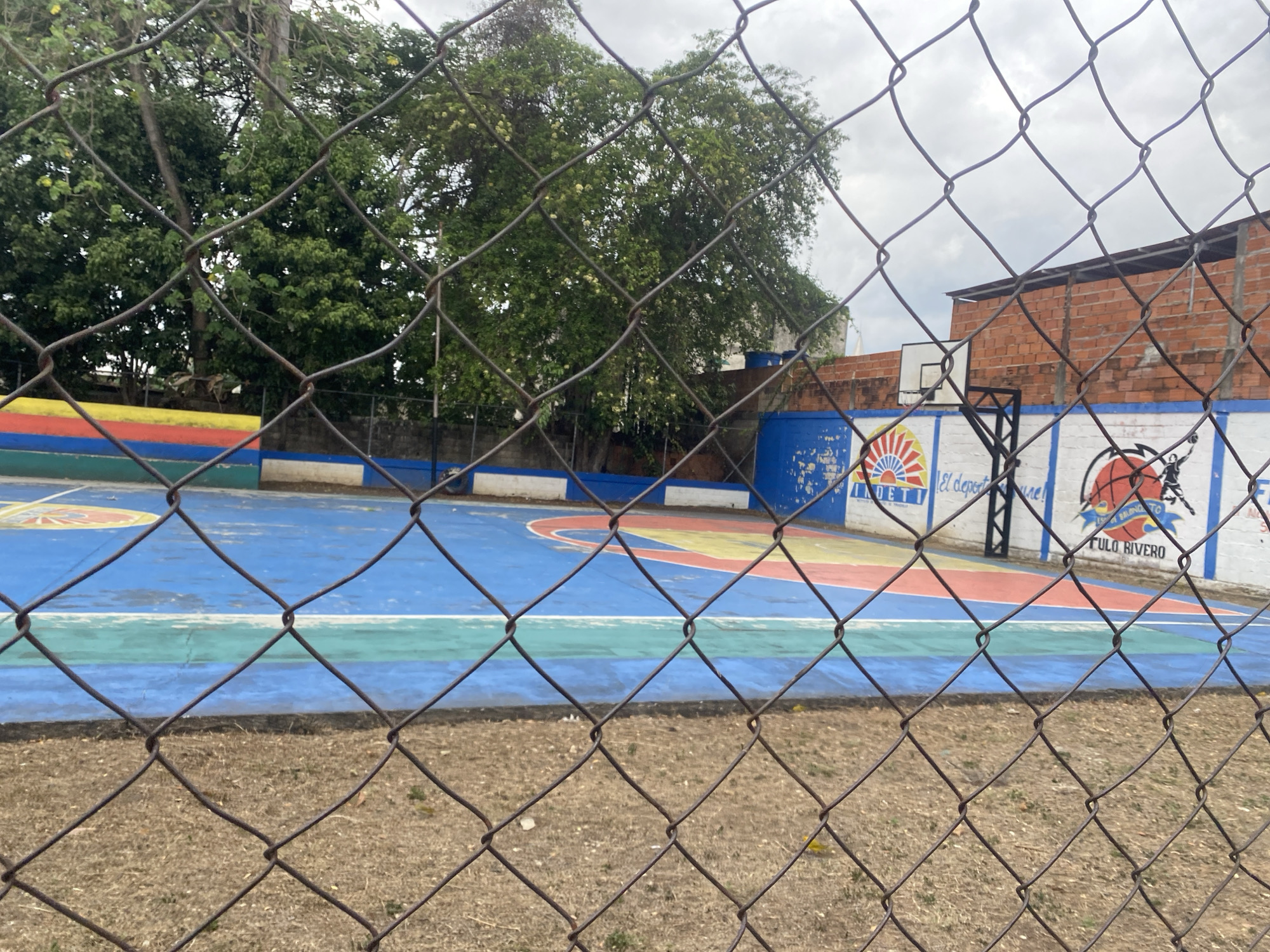 A chain-link fence obscures the view of an outdoor basketball court. The court has a blue base color with sections painted in other colors including red, yellow, and green. A backboard and basketball hoop are visible against a white wall with painted murals, including a logo that includes the letters "INDETI". A red brick building stands to the right of the basketball hoop. The surroundings appear to be an open area with dry, brown grass. Trees and a cloudy sky are visible in the background. This location is in Tinaquillo, Venezuela.