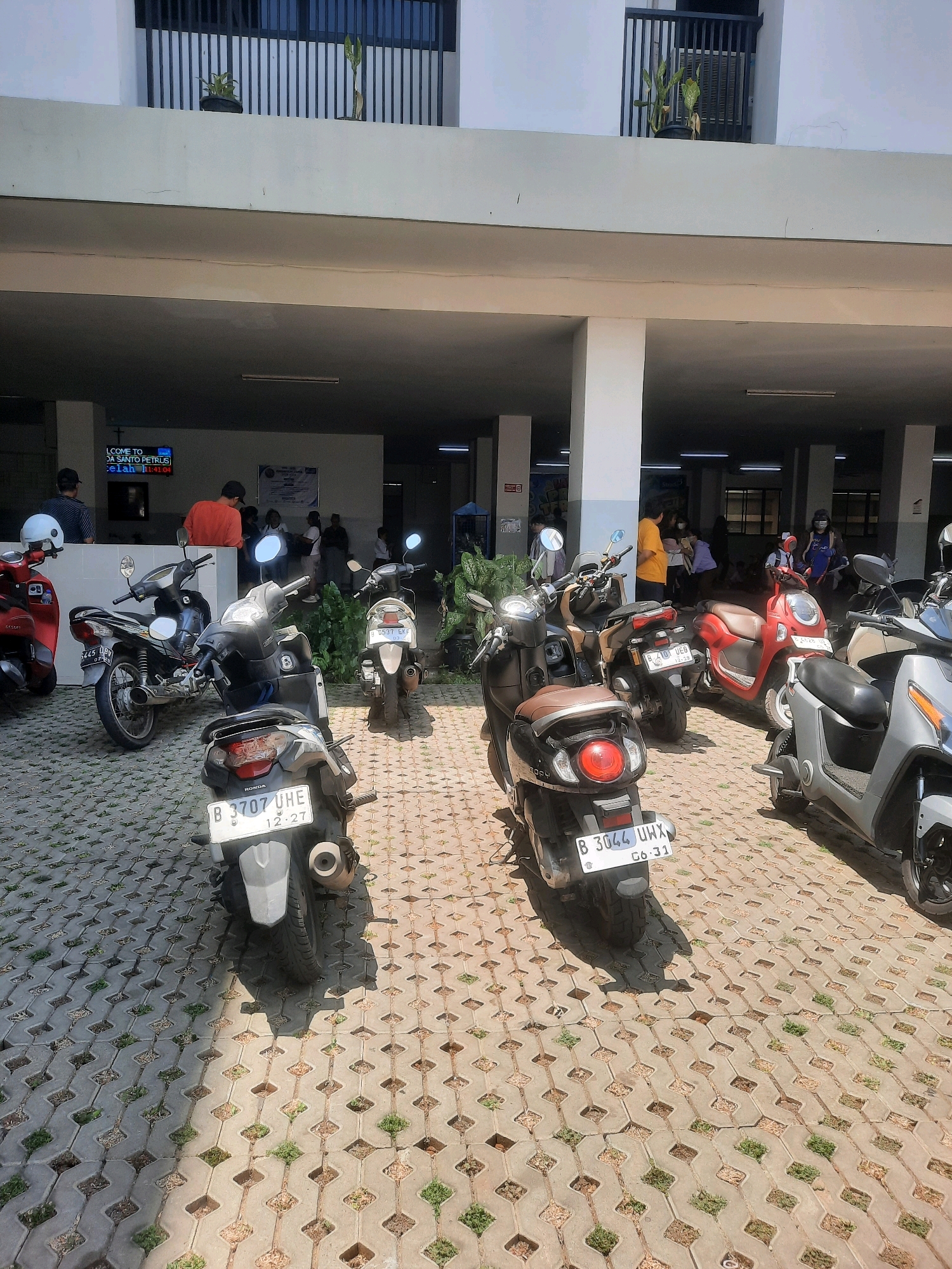 In a covered outdoor area with interlocking concrete pavers, multiple motorcycles are parked. A digital sign visible in the background reads "WELCOME TO... DA SANTO PETRUS" and displays a time. Several people are present, some standing and others appearing to be walking or interacting. The architecture suggests a public building, possibly a school or a commercial establishment. The environment is urban, consistent with a location in North Jakarta, Indonesia. The overall scene depicts a common urban setting with parked vehicles and pedestrian activity.
