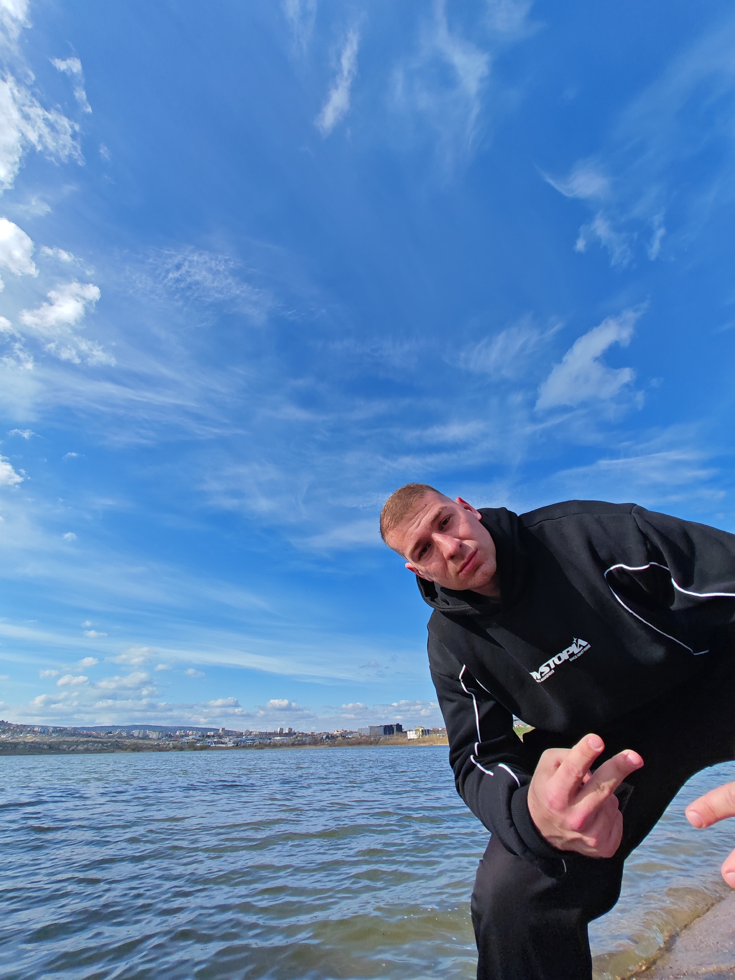 A man is photographed near a body of water with a city skyline in the background. He is wearing a black hoodie with white piping and black pants. His left hand is raised, displaying the middle finger. The skyline consists of various buildings and structures. The sky is blue with scattered white clouds. The location is near the water in the city of Shumen, Bulgaria.