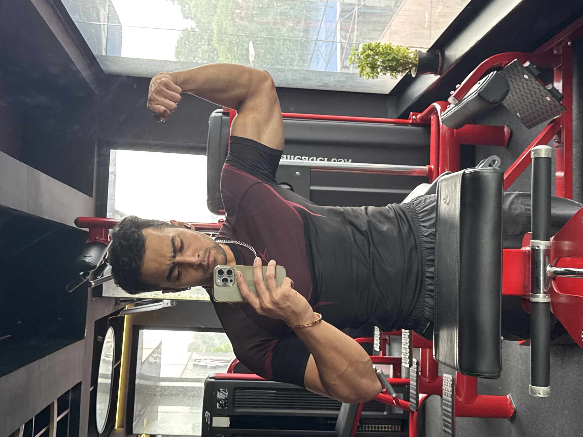 The image is a mirror selfie taken in a gym. A man is standing in front of a gym machine, wearing a maroon and black t-shirt and grey shorts. He is flexing his bicep and holding a smartphone in his other hand, which is pointed towards the mirror to take the photo. The gym appears to be modern, with red and black equipment visible. Through a large window behind him, trees and a cloudy sky are visible, suggesting the image was taken during the daytime. The context of Kolkata, India is not discernible from the image itself; no specific landmarks or elements indicate this location.