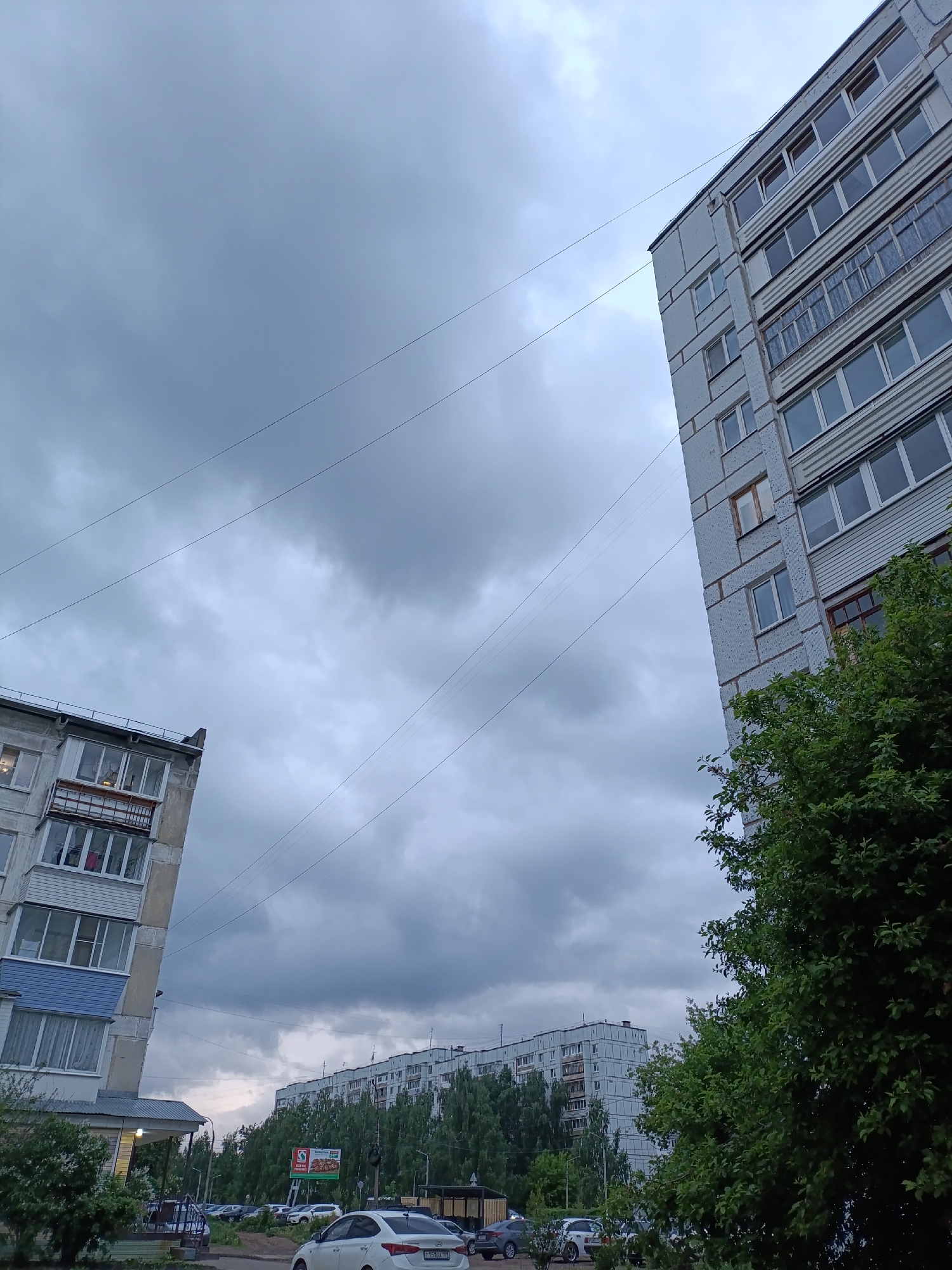 The image depicts a street scene in Chaikovsky, Russia, characterized by overcast skies. Two Soviet-era apartment buildings flank the view, with electrical lines stretched between them. In the foreground, parked cars suggest a residential area. Trees partially obscure a building further down the street, and a billboard is visible amidst the greenery. No people are directly visible in the frame.