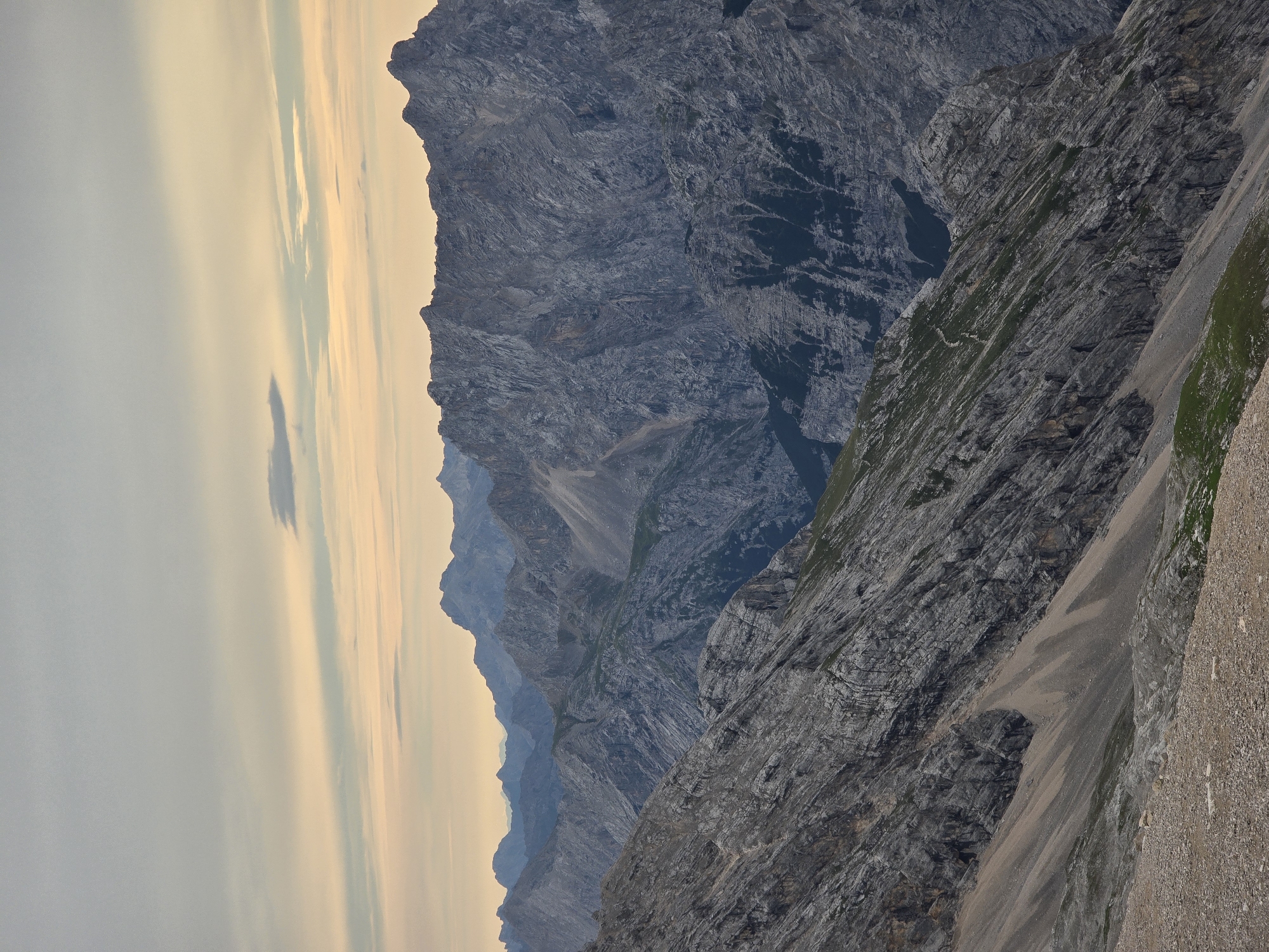The image shows a mountainous landscape with rugged, gray rock formations and patches of green vegetation. The sky above is overcast with soft, pale colors, suggesting either sunrise or sunset. The terrain is steep and appears to be a challenging hiking area. The scene is devoid of any human presence or man-made structures. The location is identified as a mountain range near Garmisch-Partenkirchen, Germany, implying it is a natural, outdoor environment.