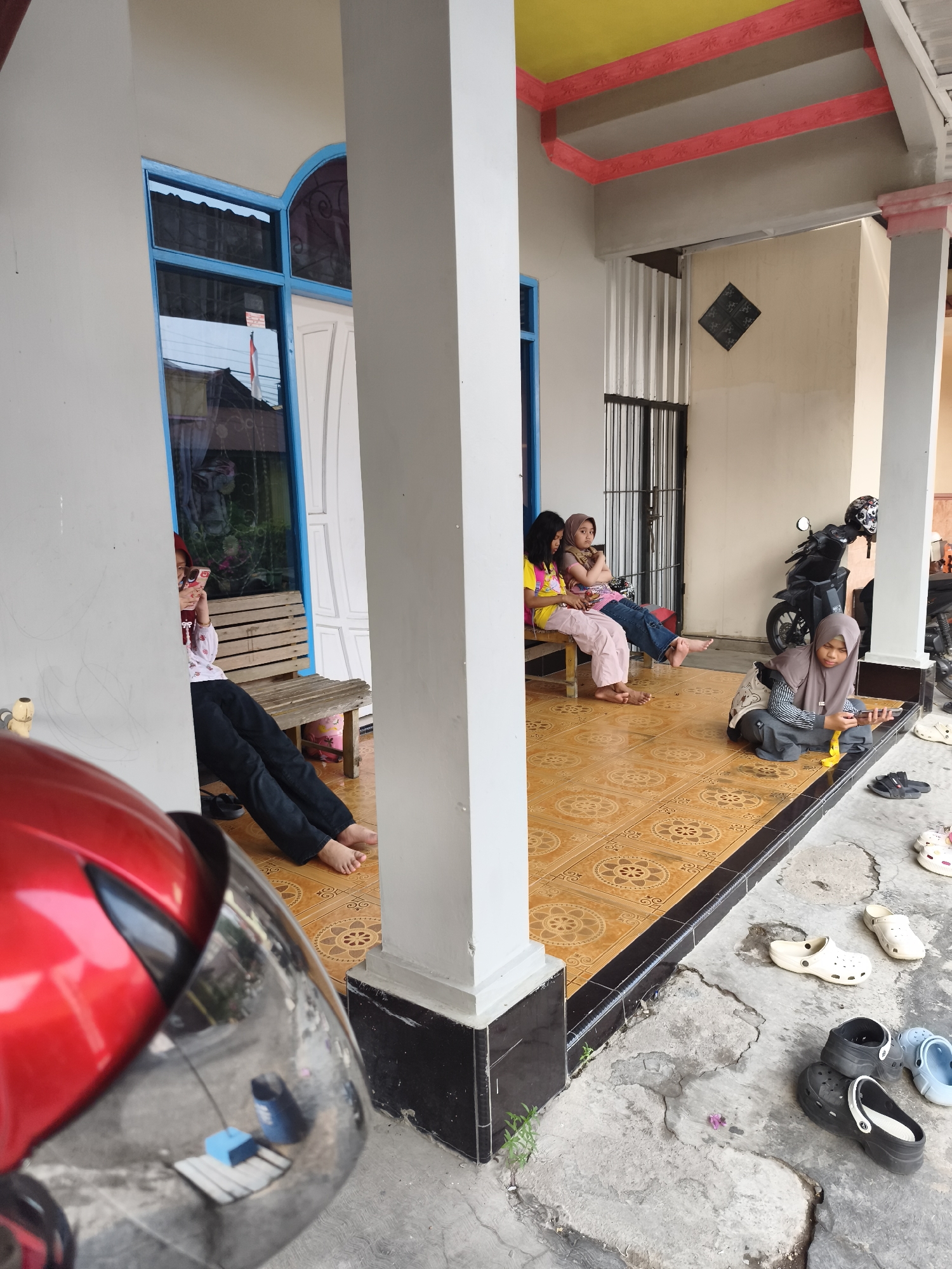 The image depicts an outdoor scene in front of a building, likely in the city of Tanjung Redeb, Indonesia, based on contextual information. In the foreground, a portion of a red motorcycle helmet is visible. The main area of focus is a tiled porch with a yellow and red decorative border on the ceiling. Several individuals are present on the porch and surrounding concrete ground.
On a wooden bench on the left, a person wearing a headscarf and a white patterned top is seated, looking at their phone. To their right, three young individuals, two girls and one child being held by one of them, are seated on a bench. They are dressed casually, with one girl wearing a yellow t-shirt and pink pants, another in a pink t-shirt and jeans, and the third, the child, is wearing a pink top.
In the center right, a person wearing a grey headscarf and grey outfit is crouched on the tiled floor, holding a smartphone. A black motorcycle with a helmet on its seat is parked behind them. Scattered on the concrete ground around the porch are various pairs of casual footwear, predominantly clogs in black, white, and blue. The environment appears to be a residential or public area with a building facade featuring a blue-framed window and a white door.