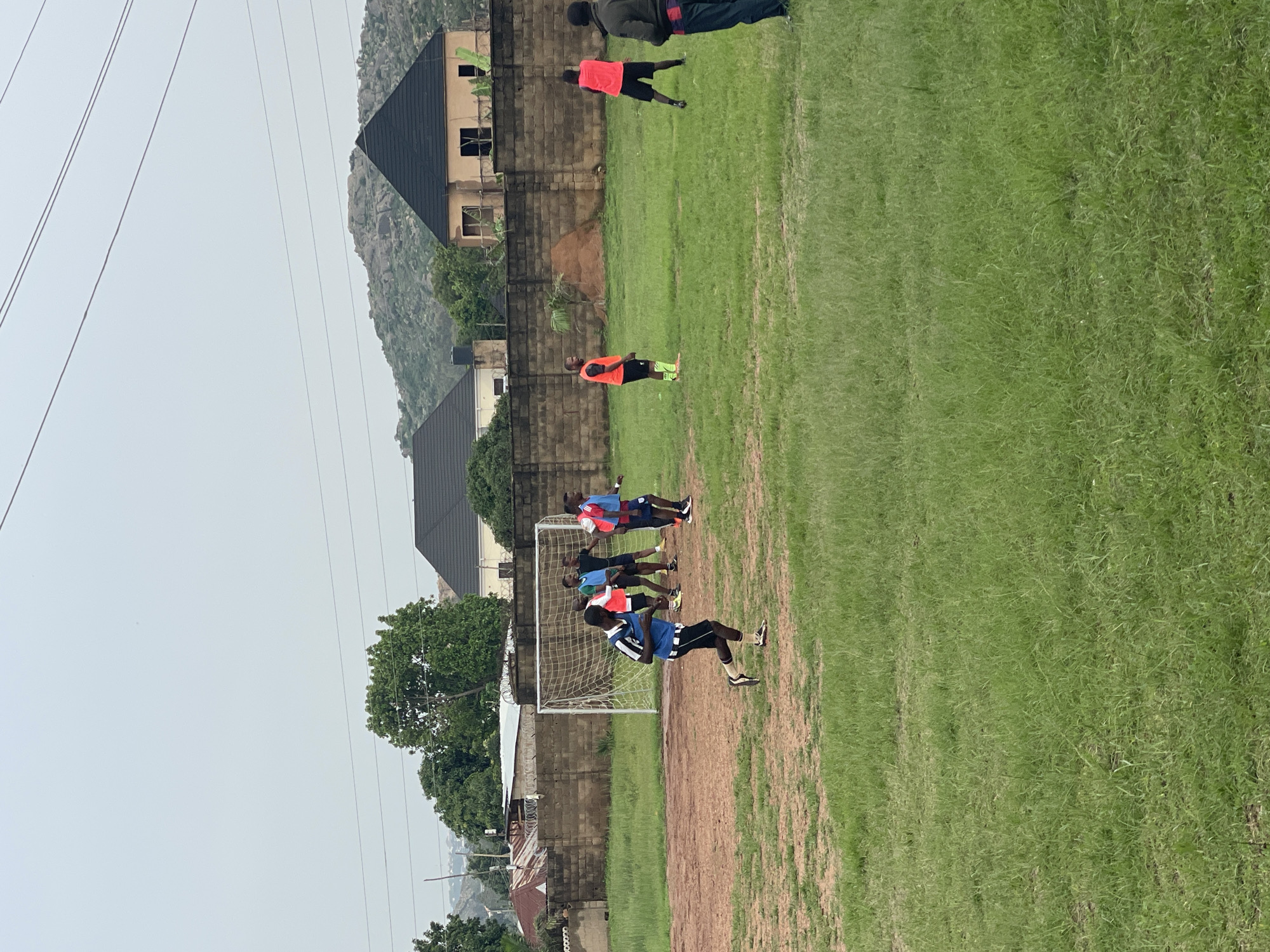 The image depicts a group of young men engaged in a soccer drill on a grassy field. The scene is set in Jos, Nigeria, with a backdrop of buildings and rocky hills. The overcast sky suggests it might be a cloudy day, possibly late afternoon or early morning.
The men are wearing athletic attire, including brightly colored jerseys and shorts. Some are in motion, while others are standing and observing. One man in a blue and white striped jersey is in the foreground, appearing to be mid-stride or performing an action near a soccer goal. Other players are positioned around the goal, some wearing red or blue vests. The soccer goal itself is visible, made of white metal with a net. The field has patches of dirt and worn grass, indicating regular use.
In the background, several buildings with dark-tiled roofs are visible. There are also trees and dense vegetation, characteristic of the Nigerian landscape. Power lines stretch across the sky, indicating the presence of infrastructure. The overall atmosphere seems to be one of training and athletic practice. There is no visible text in the image.