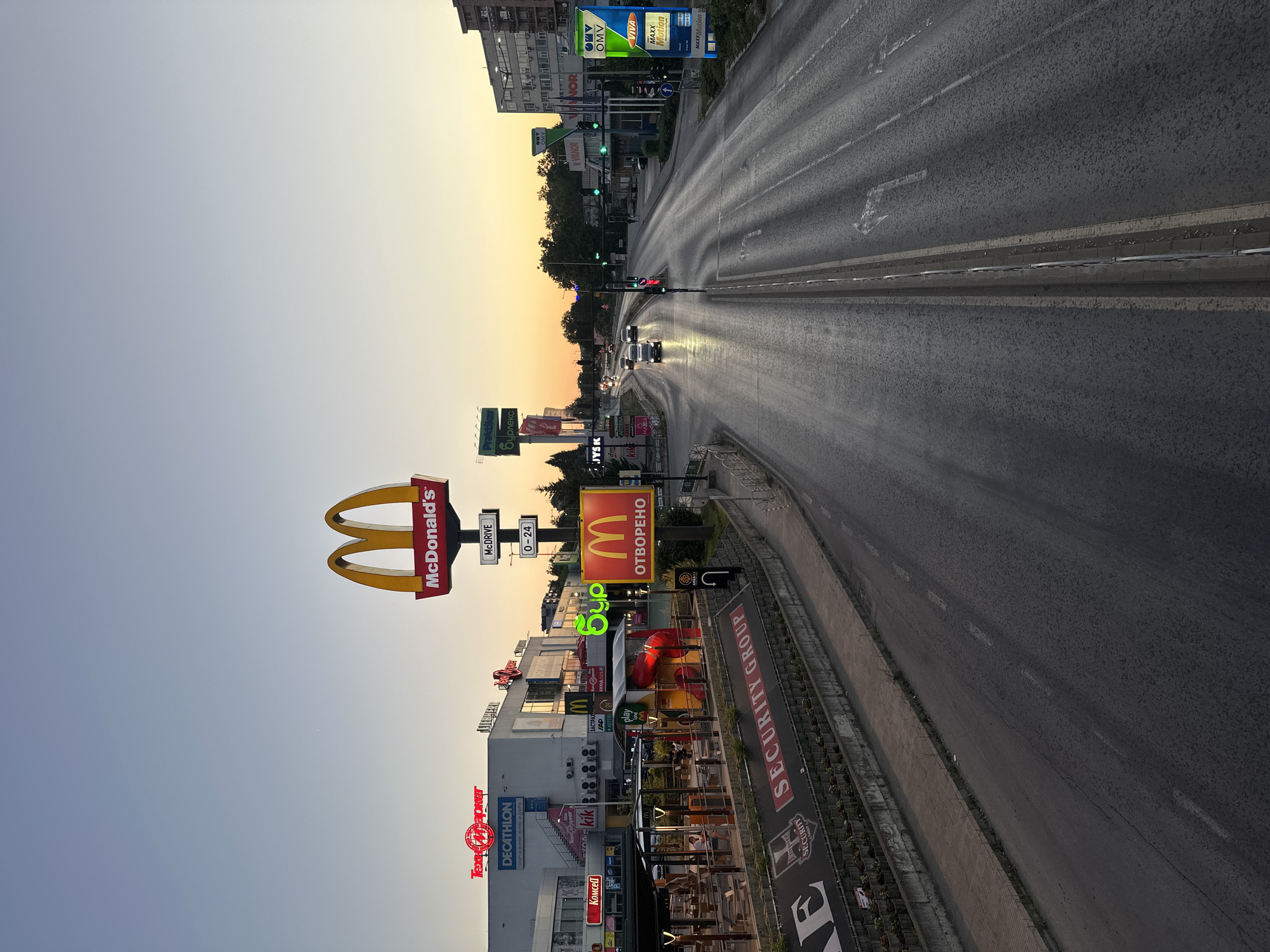 The image captures a street scene in Veliko Tarnovo, Bulgaria, during dusk. A multi-lane road dominates the foreground and middle ground, with vehicles moving in the distance. On the left side of the road, a complex of commercial buildings is visible, featuring prominent signage for McDonald's, Decathlon, and other businesses. The McDonald's restaurant is clearly identifiable by its iconic golden arches logo and illuminated sign that reads "McDonald's" and "McDRIVE". Adjacent to it is a brightly lit outdoor play area for children. Further down the street on the right, a gas station with OMV branding and a VIVA MAXx Motion advertisement is situated. The sky above displays a gradient of soft, warm colors, transitioning from yellow and orange near the horizon to a pale blue overhead, indicative of sunset. The overall environment suggests an urban or suburban commercial area, likely on a main thoroughfare. No specific individuals are clearly identifiable in the scene.