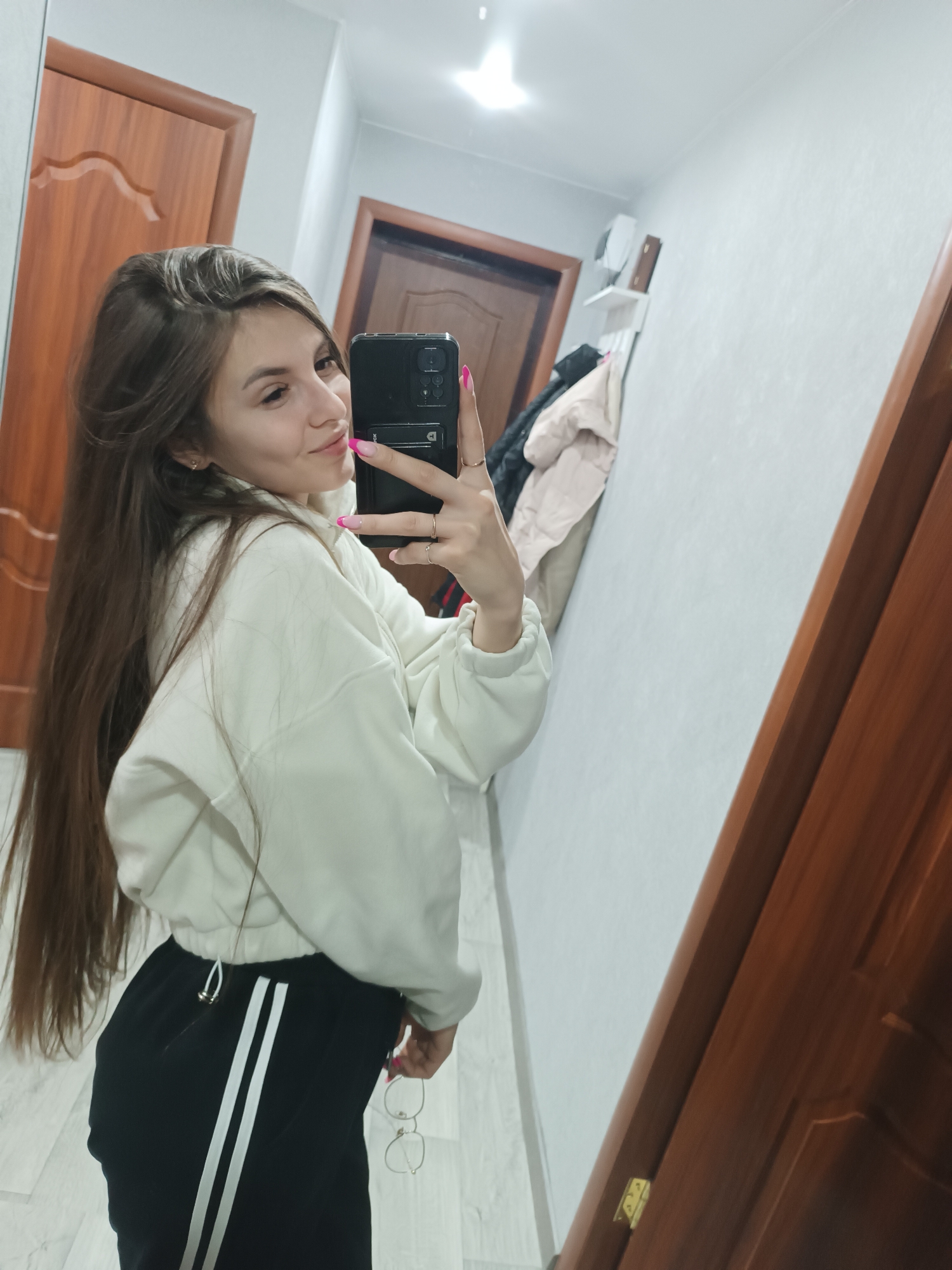 An image captures a young female taking a mirror selfie in an interior setting, likely a hallway or room, within the city of Unknown, Unknown. The subject, positioned off-center to the left, is observed from the waist up, facing towards her reflection. She has long, straight, dark brown hair and is wearing a light-colored, possibly white, long-sleeved cropped sweatshirt and black track pants featuring two parallel white stripes down the outer seams. Her fingernails are painted pink with white tips, and she wears multiple rings on her right hand. She holds a dark-colored smartphone with a visible multi-lens camera module in her right hand, extended towards the mirror to capture the reflection, and a pair of clear-framed glasses in her left hand, positioned near her hip. She is looking towards the phone/mirror with a slight smile.
The reflected background reveals light-colored, possibly white, walls. To the subject's right (viewer's left), a dark brown wooden door with a panel design is visible. Above and behind her, a wall-mounted white shelf with a small dark object on it, and two jackets (one light, one dark) hanging below, are present. To her left (viewer's right), a portion of another dark brown wooden door frame is visible, including a brass-colored door hinge at the bottom. The floor appears to be light-colored tile or laminate. An overhead light fixture reflects in the upper center of the mirror.