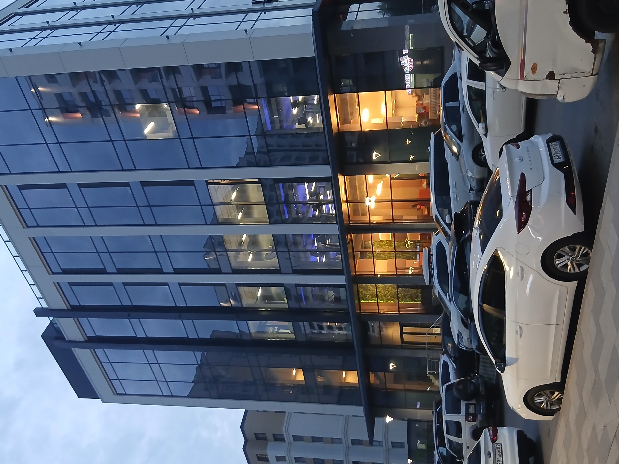 The image depicts a street scene in front of a modern glass-fronted building, likely an office building or a commercial complex. The building's facade is characterized by large, reflective glass panels interspersed with sections that glow with warm, artificial light from within. The lights suggest it is evening or night, or that the interior lights are on during the day. Several cars are parked in a row along the building's exterior, most of them white. One car, in the foreground, is a white sedan with a license plate that is partially visible but unreadable. Another white vehicle, a van or SUV, is parked behind it. The sky appears overcast, suggesting it might be evening or a cloudy day. The perspective is from a low angle, looking upwards towards the building, and the image is rotated, making the building appear to be on the left and the cars on the right. Reflections in the glass of the building show interior lights and possibly other parts of the building itself. There is a sidewalk made of paving stones in front of the parked cars. A sign with illuminated text is visible on the building's glass facade, but it is too blurry to read. The overall impression is one of a contemporary urban environment.