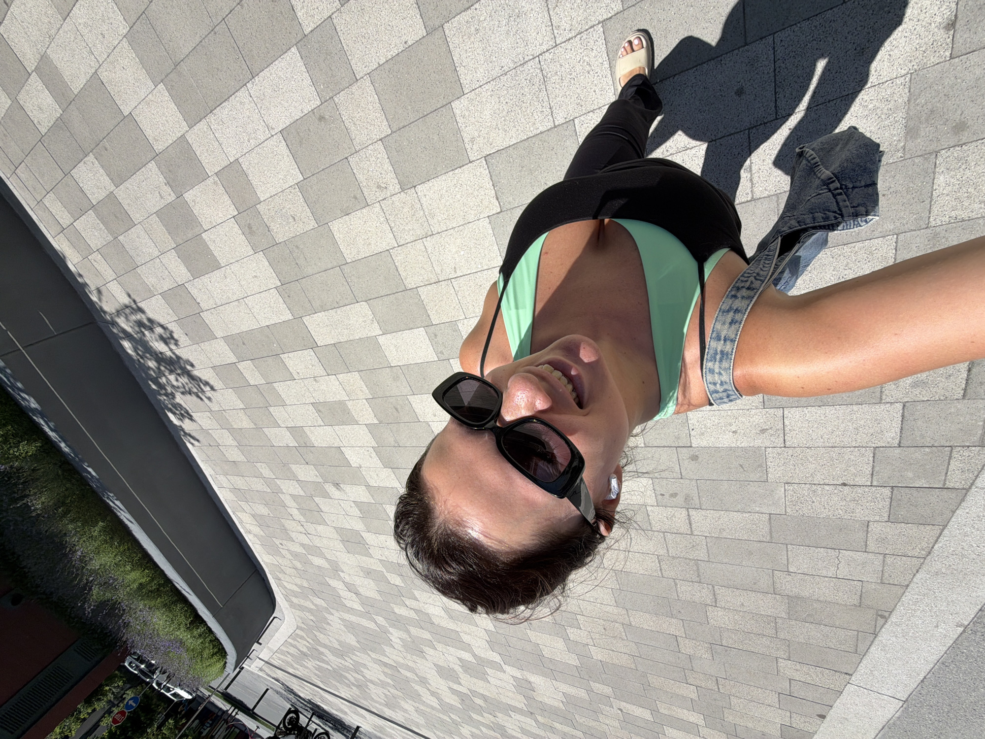 This is a high-angle, first-person perspective photo taken outdoors on a sunny day. The primary subject is a woman, seen from the chest up, looking towards the camera with a wide smile. She is wearing a light green tank top, black pants, white sandals, and large black sunglasses. She also appears to be wearing white earbuds. A denim jacket is draped over her right shoulder.
The setting is a paved walkway made of light grey and darker grey rectangular tiles arranged in a repeating pattern. To the left of the walkway, there is a street with some parked cars and a strip of green vegetation with purple flowers. The lighting suggests it is daytime, with strong sunlight casting distinct shadows on the pavement, including the woman's shadow and the shadow of tree branches.
The overall mood is cheerful and energetic, conveyed by the woman's bright smile and the sunny weather. The location context provided indicates this is in Belgrade, Serbia. No visible text is discernible in the image.