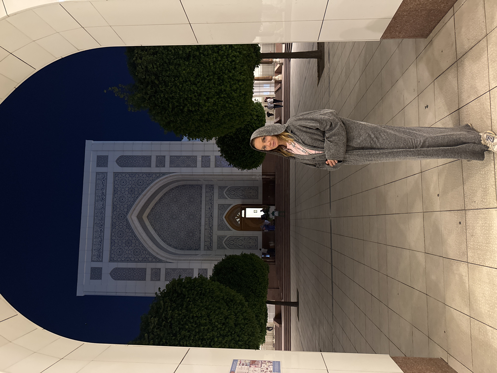 The image captures a scene at night in Tashkent, Uzbekistan. A woman stands prominently on the right, wearing a gray hooded tracksuit and white sneakers, positioned on a tiled walkway. In the background, framed by an arched white structure, is the ornate facade of the Minor Mosque, showcasing intricate geometric patterns and an arched entrance. Two neatly trimmed trees flank the walkway leading to the mosque, adding depth to the composition. A few individuals are vaguely visible near the mosque's entrance. The sky above the mosque is a deep blue, suggesting it is past dusk.