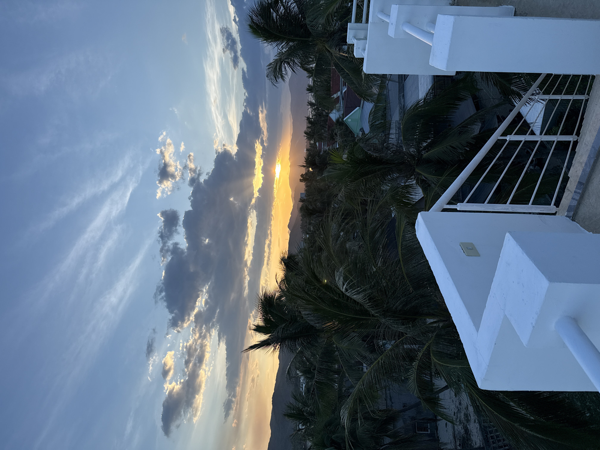 A view from a rooftop overlooking palm trees and a town at sunset. The sky is a mix of blue and orange with scattered clouds. The sun is low on the horizon, casting rays of light through the clouds. In the foreground, there is a white railing and steps. Palm trees are visible throughout the midground, with rooftops of houses and buildings peeking through them. In the distance, mountains form a silhouette against the colorful sky. The overall impression is of a tropical or coastal location at the end of the day.