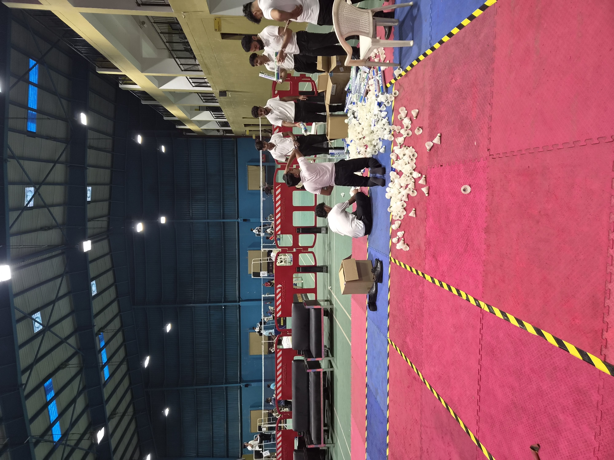 This image depicts an indoor badminton court located in Phagwara, India. Several young men, dressed in white polo shirts and black pants, are gathered on the court floor, surrounded by numerous badminton shuttlecocks. Some individuals are holding shuttlecock tubes, while others are kneeling or standing amongst the scattered shuttlecocks. Boxes and a white plastic chair are also visible in the foreground. In the background, more badminton courts are set up with nets and seating areas, and several other people can be seen in the distance. The environment appears to be a sports facility with a high ceiling, bright artificial lighting, and blue and yellow taped markings on the court floor.