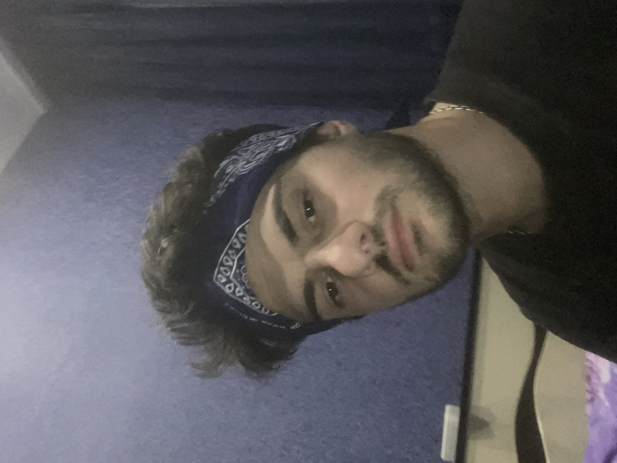 A male individual, appearing to be in his early to mid-twenties, is captured in a close-up, front-facing selfie. He has dark, tousled hair and is wearing a blue bandana with a white paisley pattern. His facial features include prominent eyebrows, dark eyes, a straight nose, and a short beard. He is wearing a black t-shirt and a silver chain necklace. The background is a textured, purple wall, with a dark blue curtain visible to the right. To the left and below, a portion of a bed with patterned bedding and a dresser with a white object on it can be seen. The lighting suggests an indoor setting, likely a bedroom, with the source of light appearing to be from the front and slightly above the subject. The overall composition is typical of a casual self-portrait. The image does not provide any specific visual cues to determine the location within Tbilisi, Georgia.
