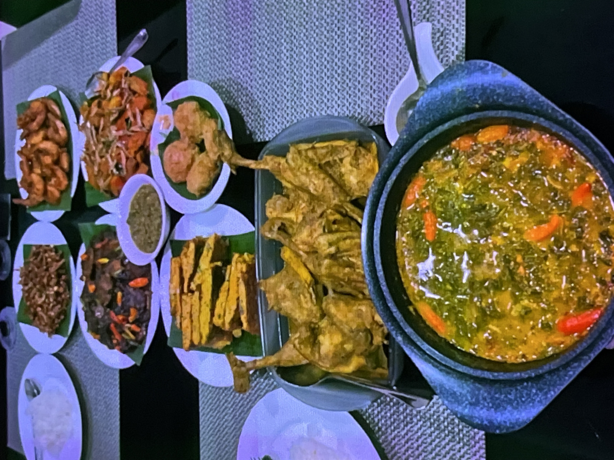 The image depicts a lavish spread of Indonesian cuisine laid out on a table. The dishes include rice, fried anchovies, a dark meat dish with chili peppers, shrimp, a salad-like dish, fried tofu, fried chicken pieces, and a bowl of soup. The food is presented on white plates and in a large dark serving dish. The setting appears to be an indoor dining area, possibly a restaurant or home in Cadek, Indonesia, given the style of the food and presentation. There are no people visible in the image. The overall impression is one of a festive or celebratory meal.