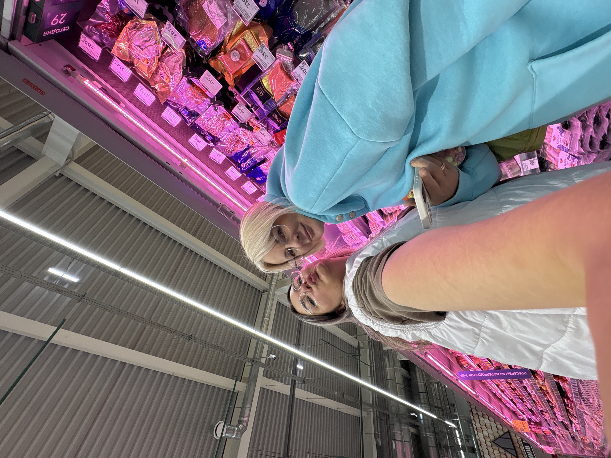 Here is a forensic description of the image:
**Scene:** The photograph shows two individuals, likely women, taking a selfie in what appears to be a supermarket. The location is implied to be in Birobidzhan, Russia based on the text visible in the image.
**People:**
* Two people visible, presumably women. One has short blonde hair and is wearing glasses and a light blue sweatshirt. The other has dark hair and glasses, and wears a white vest.
**Subjects & Objects:**
* Shelves of pre-packaged meat products are present in the background with pink lighting.
* Product labels and pricing are visible, with Russian text.
* The ceiling of the supermarket is visible, showing metal beams and lighting fixtures.
**Actions:**
* The two women are holding a phone to take a selfie, looking into the camera.
**Interactions:**
* The women are posing together, likely indicating a friendly or social interaction.
* The women are interacting with the supermarket environment by being present near the products and taking a photo.
**Environment:**
* The setting is a supermarket aisle with meat products displayed for sale.
* The presence of Russian text indicates the scene is in Russia.
* Overall lighting is a combination of fluorescent and pink lighting on the shelves.