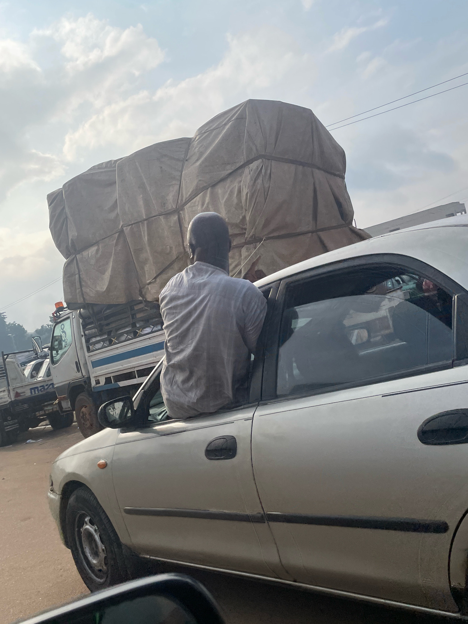 A man, viewed from behind, is sitting on the exterior of the open driver's side window of a silver sedan, his torso and head extending outwards. He is wearing a light-colored, possibly grey, short-sleeved shirt. The sedan is in traffic, with a white truck carrying a large, tarp-covered load of goods to its left. The truck has a blue stripe along its side and an orange flashing light on its roof. Another vehicle is partially visible behind the truck. The scene appears to be on a road in Abuja, Nigeria, with overhead wires visible against a cloudy sky. The road surface is reddish-brown.
