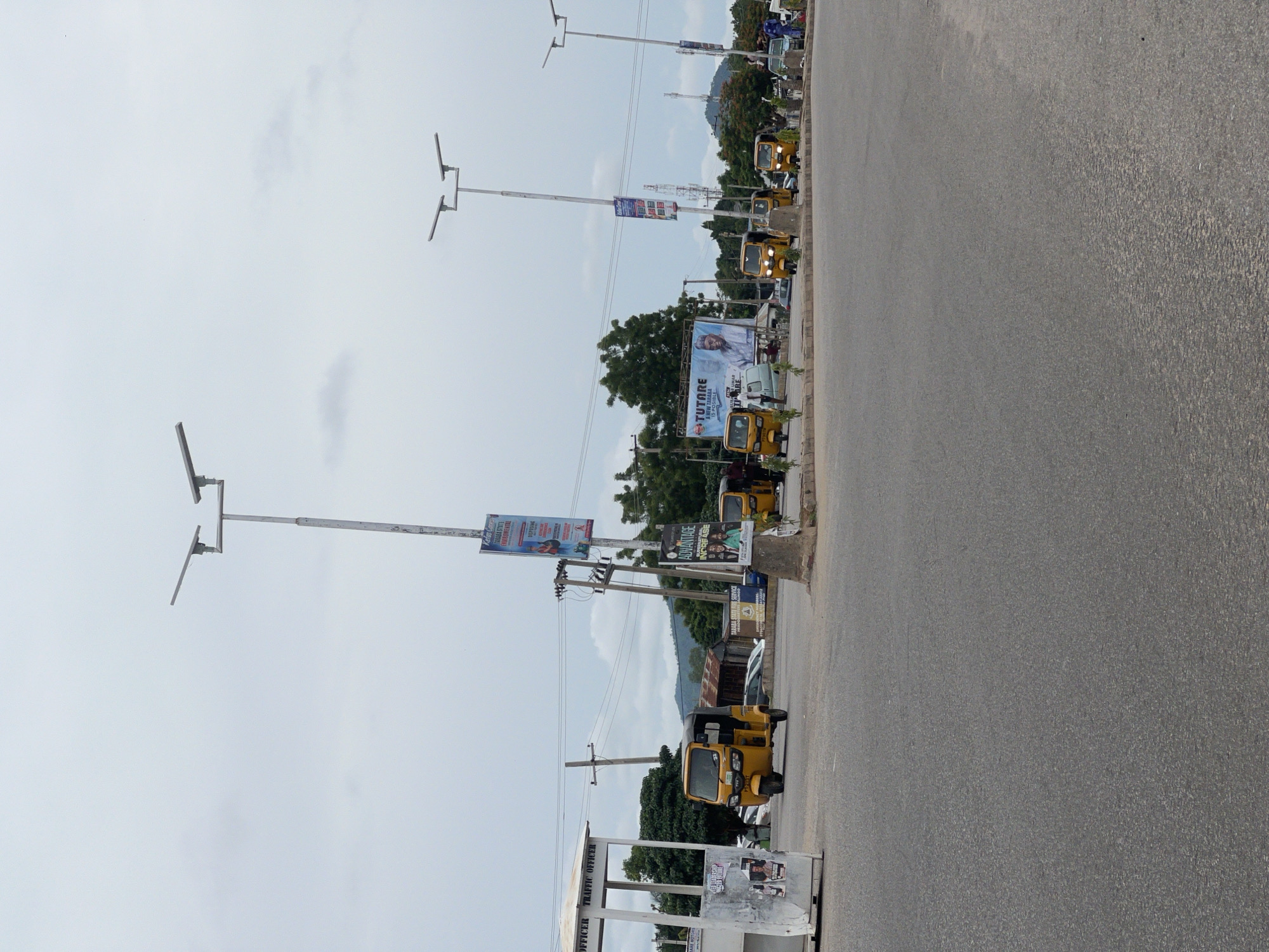 The image depicts a roadside scene in Jalingo, Nigeria. The primary subjects are several yellow auto-rickshaws, commonly known as Keke Napep, parked along the side of a paved road. Two streetlights with modern LED fixtures are prominently visible, casting their illumination. In the background, lush green trees suggest a possibly rural or semi-urban setting, with a distant view of hills. The sky is overcast, indicating a cloudy day, and the lighting suggests it is daytime.
Several large billboards and signs are posted along the roadside. One billboard features the text "TUTARE A NEW TARABA IS POSSIBLE" alongside a portrait of a man. Another sign reads "ADVANTAGE INCREASE". A smaller sign also indicates "TARABA STATE FIRE SERVICE". Near the bottom of the image, a structure with the words "OFFICER TRAFFIC OFFICER" is partially visible, suggesting a traffic management point. The overall impression is of a typical urban or peri-urban environment in Nigeria, characterized by local transportation, public advertising, and infrastructure elements. No specific activity or event is clearly depicted, beyond the general presence of vehicles and infrastructure.
