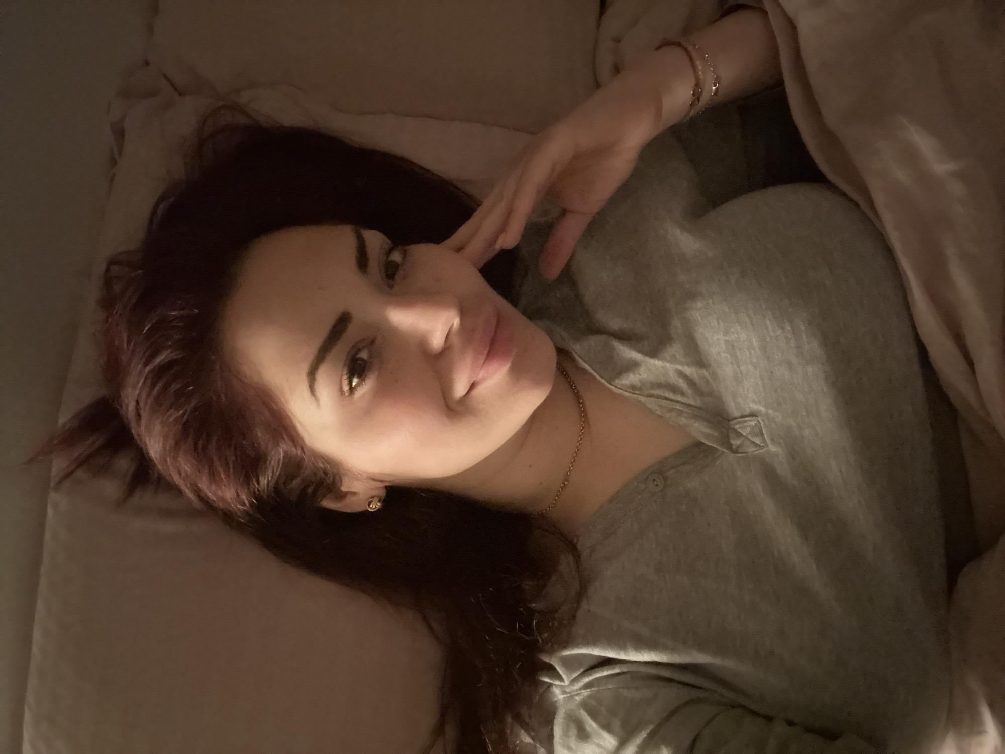 A woman is lying on a bed covered in beige bedding. She is wearing a grey button-down shirt and a gold necklace. Her right hand is touching her face. She has long, dark red hair. Location: Not possible to determine the city of Sofia, Bulgaria.