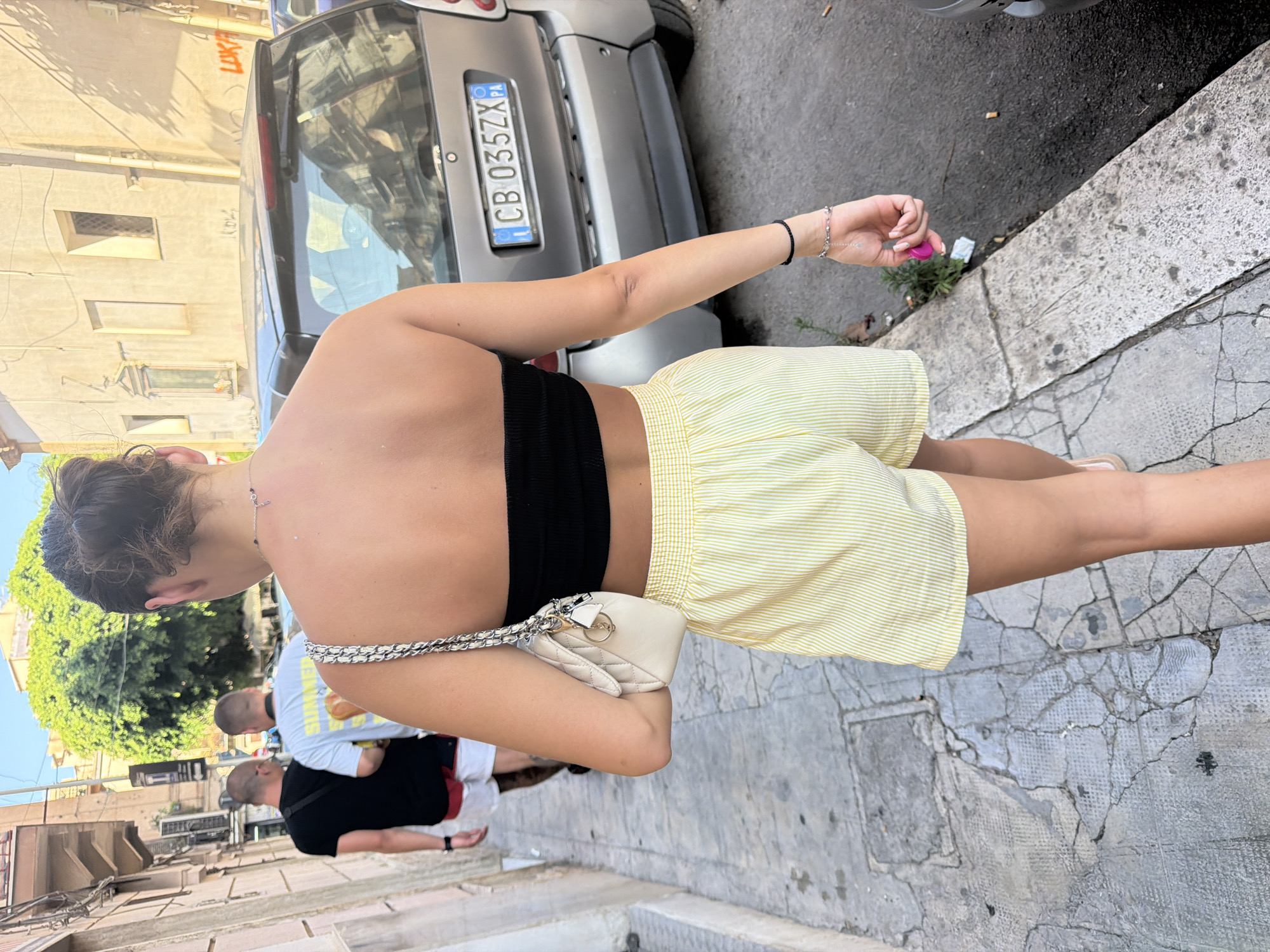 A rear view of a young woman walking down a narrow city street in Palermo, Italy. She is wearing a black bandeau top and yellow and white striped shorts. She carries a quilted beige purse on her left shoulder and a silver charm bracelet on her right wrist. Two men are walking ahead of her on the same sidewalk. The man on the left wears a black t-shirt and red and white shorts. The man on the right wears a white t-shirt with an image printed on the back and grey shorts. Behind the people, a silver Smart car is parked on the right side of the street, with a license plate reading "CB 035ZX". Buildings line the left side of the street, and the street surface is made of cracked stone pavers. The sky is clear and blue.