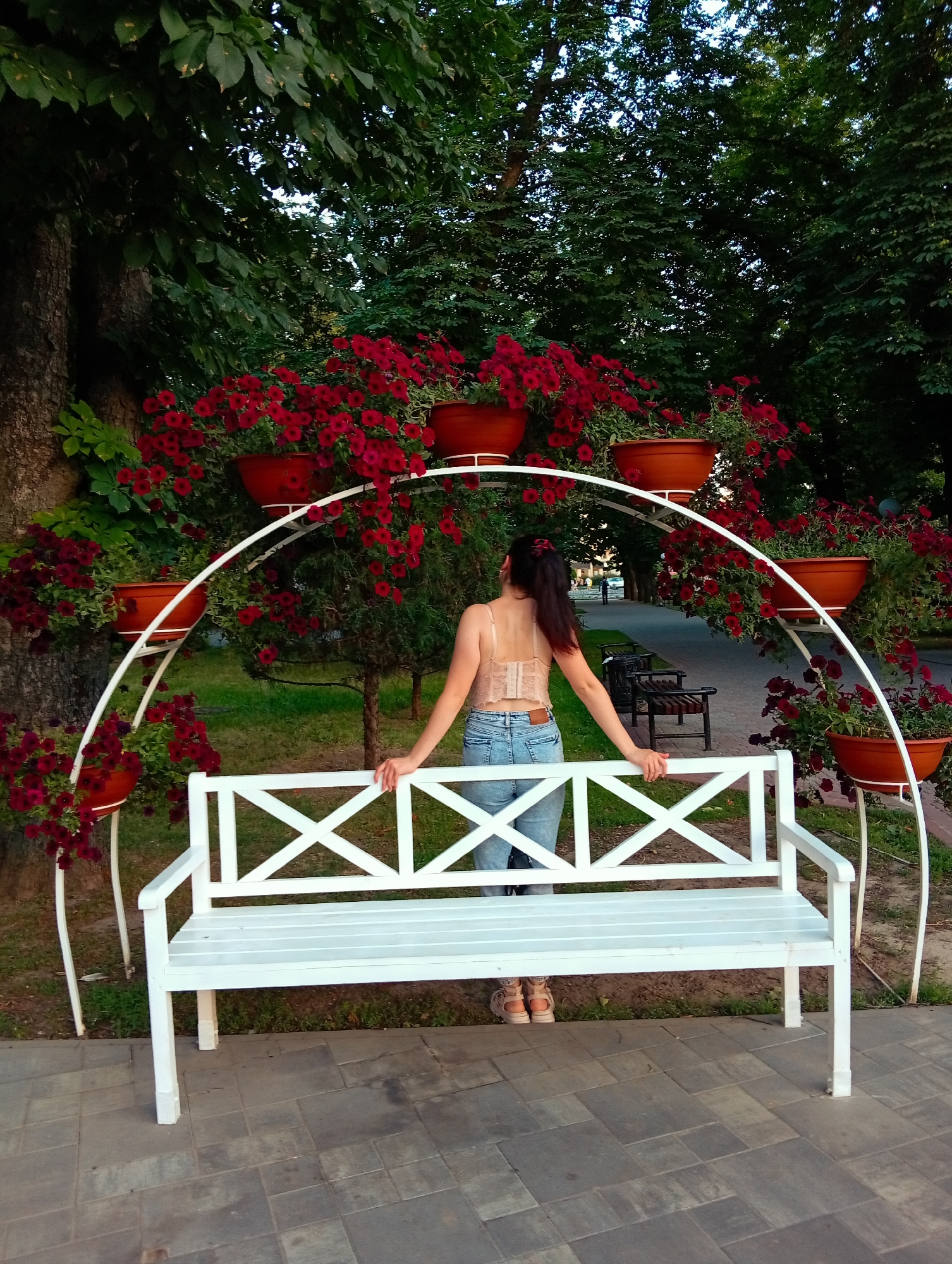 A person stands with their back to the camera, facing away from a white bench in a park. The person is wearing a light-colored lace-trimmed top and blue jeans. They are holding onto the back of the bench with both hands. The bench is positioned in front of a white arched structure with multiple shelves holding terracotta pots filled with red flowers. The scene is outdoors, with trees and greenery in the background. The ground in front of the bench is paved with gray stones. The time of day appears to be late afternoon or early evening, suggested by the soft lighting and long shadows. The overall mood is peaceful and serene. There is no visible text in the image.