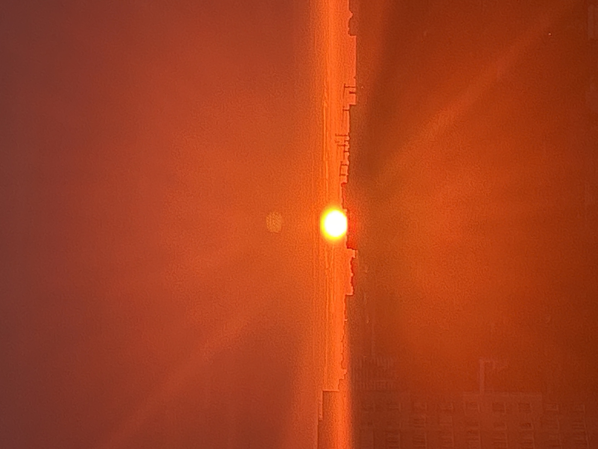 The image captures a sunrise over the city of Ivanteyevka, Russia. The sun, a bright white orb, is positioned low on the horizon, partially obscured by a faint layer of clouds or atmospheric haze, casting a warm, orange glow across the sky. In the foreground, a silhouette of the city's skyline is visible, featuring various buildings, including what appear to be apartment blocks with discernible window patterns. The overall scene is dominated by shades of orange and red, indicative of a dramatic sunrise. No people or specific actions are discernible in the image. The environment appears to be urban.