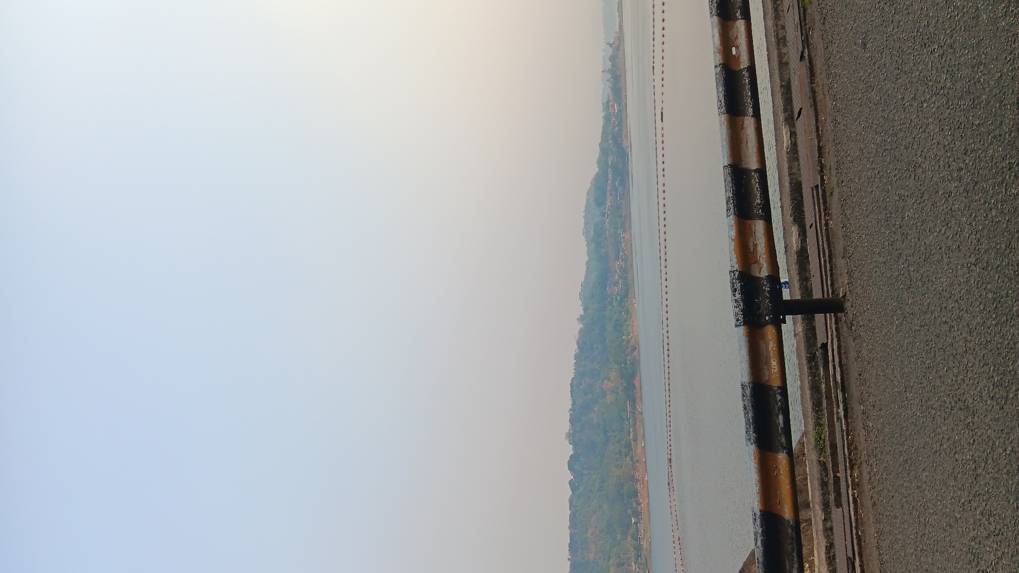 The image shows a view of Jatiluhur Reservoir in Purwakarta, Indonesia, from a road with a guardrail. The reservoir is calm and reflects the hazy sky. A line of buoys stretches across the water, possibly marking a boundary or a swimming area. The background features a green, tree-covered hillside dotted with houses, suggesting a settlement near the reservoir. The guardrail in the foreground is painted with alternating black and yellow stripes, showing signs of wear and tear. The road surface in front of the guardrail is asphalt. The overall atmosphere is tranquil and slightly overcast. There are no people visible in the image.