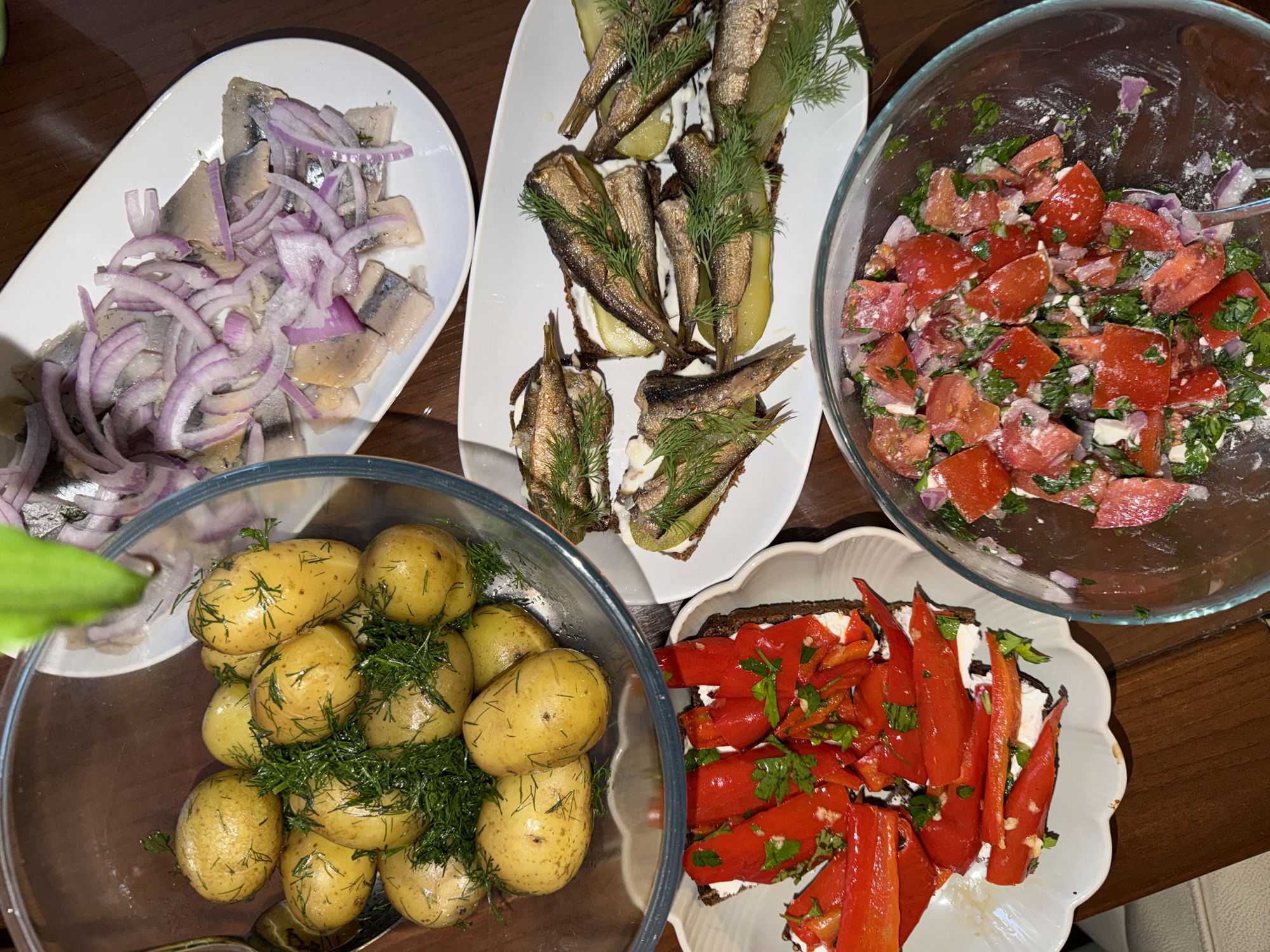 The image shows a top-down view of five dishes arranged on a wooden surface.
1. **Herring with Onion**: A white, elongated dish contains sliced herring topped with thinly sliced red onion.
2. **Sprat Sandwiches**: A similar white dish presents open-faced sandwiches. These consist of dark bread, a white spread, a pickle slice, and several small fried sprats, garnished with dill.
3. **Tomato Salad**: A large, clear glass bowl holds a salad of chopped tomatoes, onions, and herbs.
4. **Potatoes with Dill**: Another large, clear glass bowl contains cooked small potatoes, coated with butter and dill.
5. **Roasted Red Peppers**: A white, decorative dish contains slices of roasted red peppers served on bread with a white spread and parsley.
The food is presented in a casual, family-style setting. There is no indication of the location in Budva, Montenegro, nor are there people or indications of interactions visible in the image.