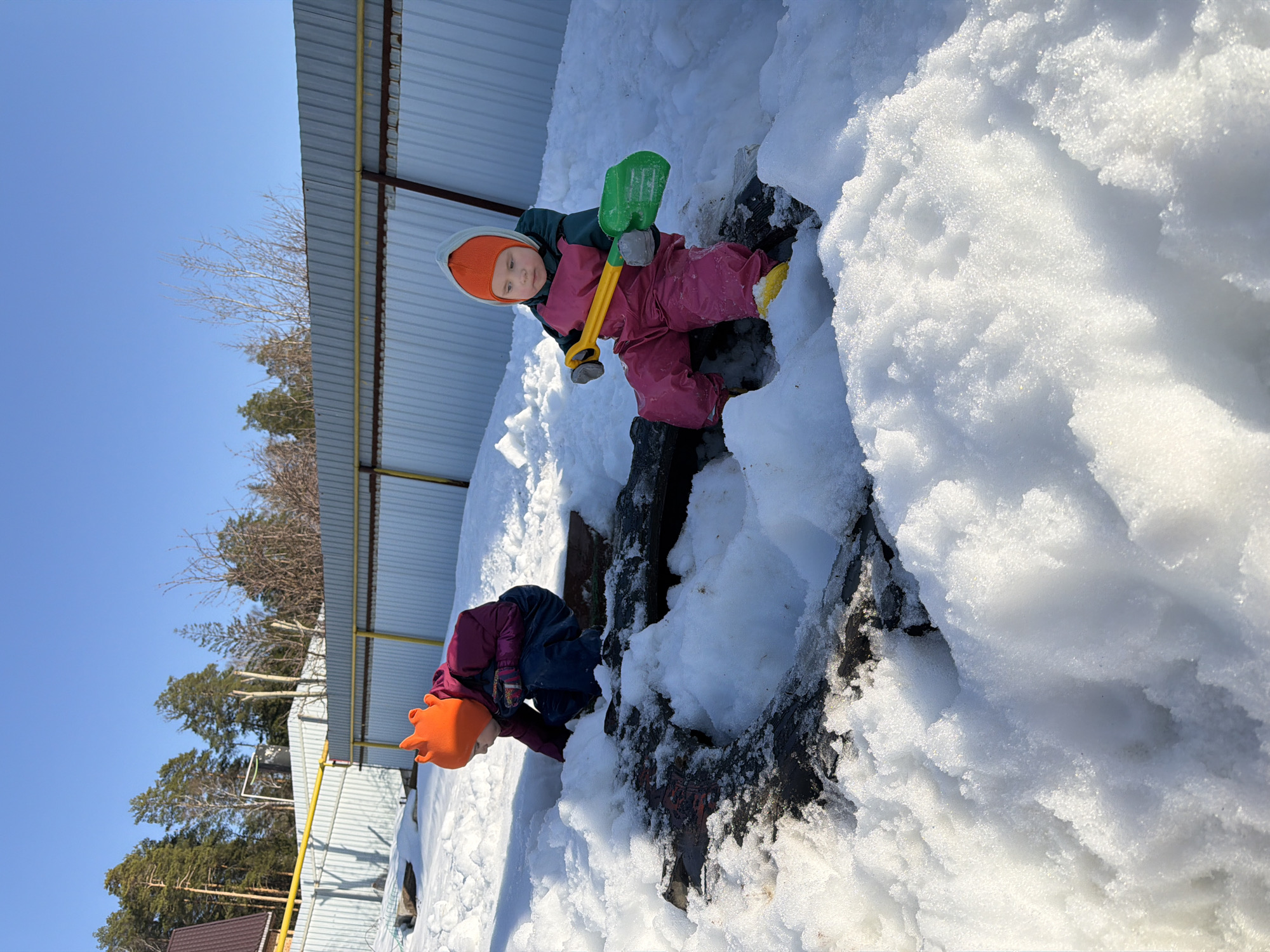 Two children are playing in the snow. One child, wearing a pink snowsuit and orange hat, stands near a large mound of snow and holds a green and yellow toy shovel. Another child, also wearing an orange hat, kneels near the snow mound. The location appears to be Chaikovsky, Russia. A metal structure with yellow supports is visible in the background, along with trees and a blue sky.