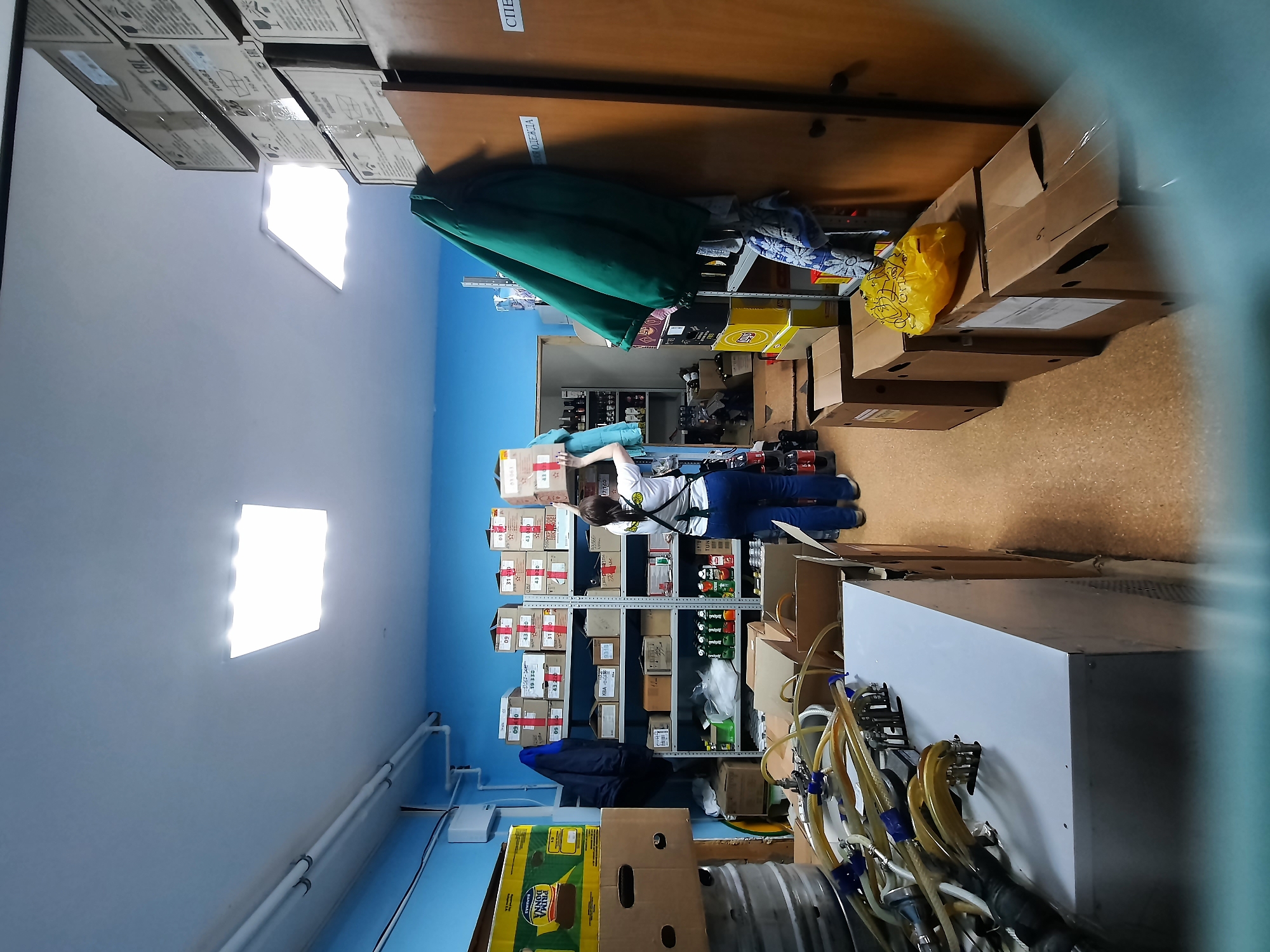 The image depicts a room in Krasnogorskoye, Russia. A woman wearing a white shirt and blue pants is standing near a metal shelving unit loaded with boxes and bottles. The unit is against a blue wall. To the right of the shelving unit are cardboard boxes and a white machine with multiple hoses. Above the shelving unit, a wooden shelf runs along the wall, with hanging clothes and more boxes on it. The room is lit by bright square ceiling lights. There are boxes stacked up on the ceiling.