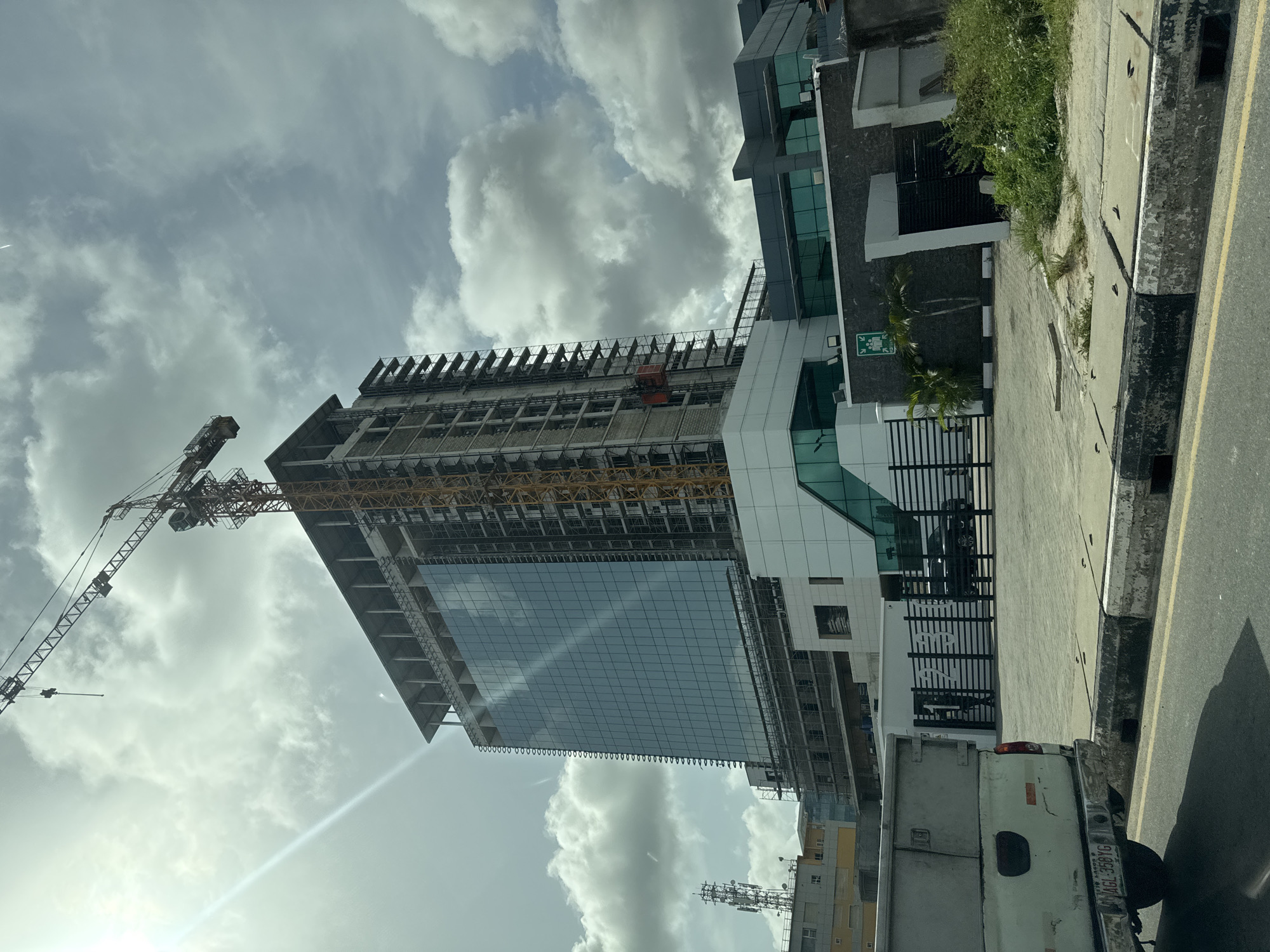 The image captures a bustling urban scene in Itirin, Nigeria, centered around a modern high-rise building under construction. The structure shows a contrast between its lower half, clad in reflective blue glass, and its upper levels, which reveal exposed concrete framework and scaffolding, indicating ongoing development. A prominent yellow construction crane towers beside the building, its arm extending across the sky, signifying active construction work.
Adjacent to the construction site is a completed multi-story building featuring contemporary architecture with dark grey paneling and green-tinted glass windows. A green emergency exit sign is visible on its exterior. At ground level, a paved road with a yellow lane marking runs alongside a concrete curb and a sidewalk. Green foliage, including bushes and small palm trees, dots the landscape along the sidewalk and within the property boundaries.
A white truck with a visible Nigerian license plate (LAGOS AGL-358YG) is parked on the roadside. Behind a black grated fence, which encloses part of the property, another dark vehicle is partially visible. A stylized "RMR" logo is seen on a white container or structure within this fenced area. The sky is largely overcast with white and grey clouds, but bright sunlight breaks through in the lower left, creating a lens flare, suggesting it is daytime.