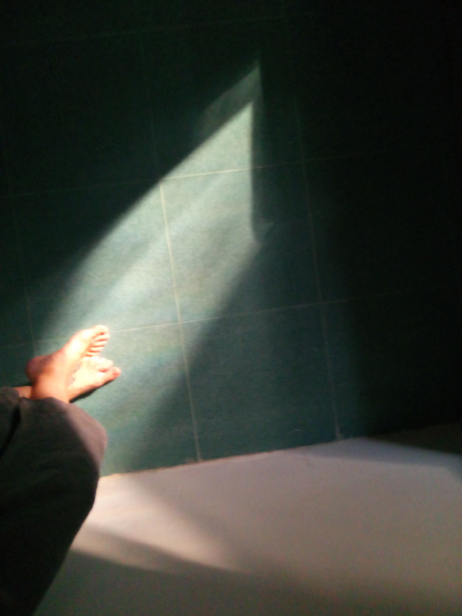 The image shows a portion of a room, possibly a bathroom, with green tiled walls and a lighter-colored floor. Sunlight streams in from an unseen source, creating a bright rectangular patch on the tiles. A person's feet are visible within the sunlit area, along with the lower part of their legs covered by dark-colored fabric. The location is unknown, and not possible to determine from this image.