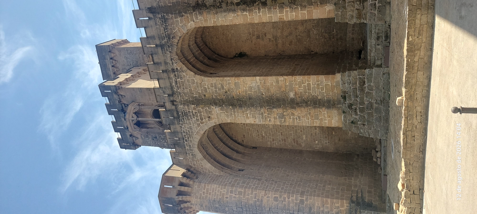 The image shows a low-angle view of the Olite Castle, also known as the Royal Palace of Olite, located in Olite, Navarre, Spain. The castle is constructed primarily of brown stone blocks, exhibiting signs of age and weathering. The prominent features are large arched openings and crenellated battlements, indicative of medieval fortress architecture. The sky above is blue with scattered wispy clouds, suggesting fair weather. The ground in the foreground is paved and appears to be an open public space, with a low stone wall and a dark metal post visible. A timestamp on the bottom left reads "12 de agosto de 2026 14:04," indicating the date and time the photograph was taken. No people are visible in the image.