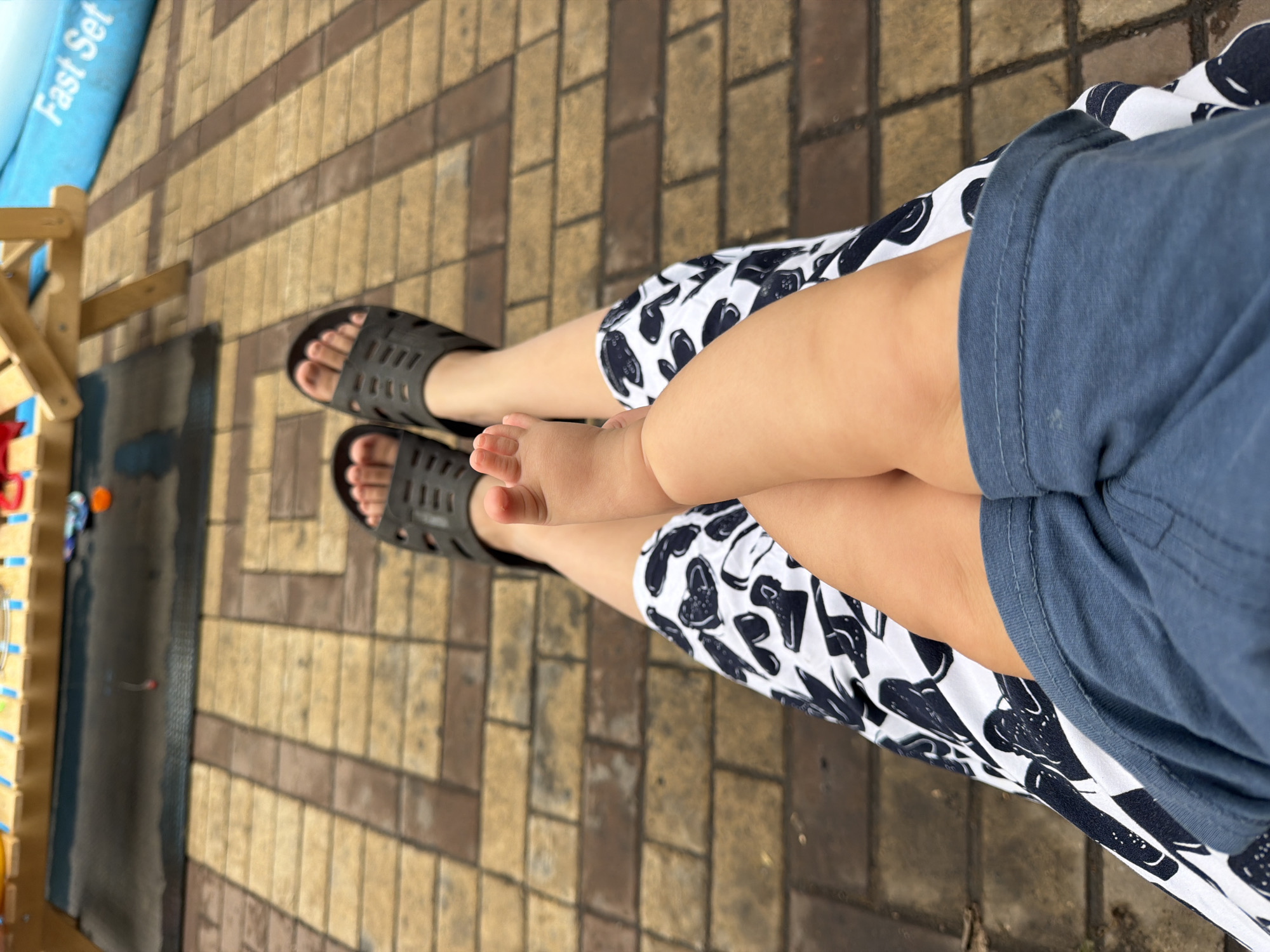 The image shows a person's legs and a baby's leg, suggesting they are sitting together on a paved patio area. The person is wearing dark grey slide sandals and a white skirt with a black pattern of what appears to be cartoon-like figures or possibly shoes. The baby's leg is bare and positioned on top of the person's leg. The background features a tiled or brick-like surface and a blue inflatable object with the text "Fast Set" visible, possibly indicating a swimming pool or water play area. The scene appears to be outdoors, potentially in a recreational setting. No specific location within Qarağandı city administration, Kazakhstan can be identified from the visual information provided.