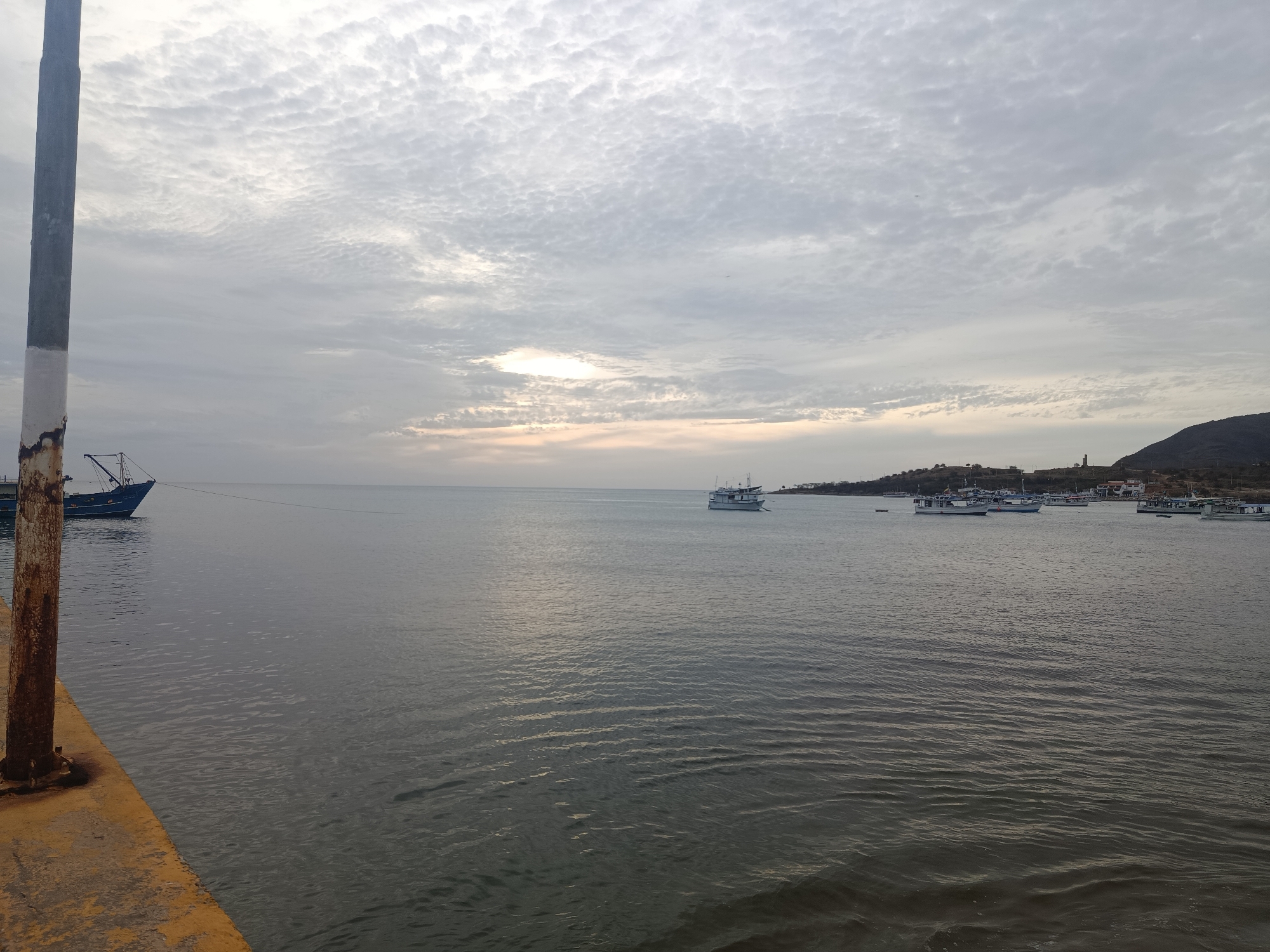 The image depicts a coastal scene in Juan Griego, Venezuela, under an overcast sky. A prominent, weathered concrete or metal pole, dark grey with a white band and visible rust at its base, occupies the left foreground, rising from a yellow-paved pier surface. The majority of the lower frame is a calm body of water, exhibiting subtle ripples across its grey surface.
On the water, several vessels are visible. A dark blue fishing vessel, possibly a trawler, with a visible crane-like apparatus, is moored or anchored in the left midground, partially behind the foreground pole. A thin line extends horizontally from this vessel across the water. Further to the right, a white multi-deck boat is anchored in the mid-distance. Along the distant shoreline to the right, a cluster of smaller white and blue boats are moored or anchored near the land.
The horizon is characterized by a dark, undulating landmass, consistent with hills, on the right side. Scattered buildings and structures are discernible along the base of these hills, including a faint tower-like structure on the far right. The sky overhead is heavily covered by a dense formation of altocumulus clouds, creating a textured, rippled pattern across the entire upper half of the image. A soft, diffused light source, likely the sun obscured by clouds, is visible as a lighter glow near the horizon in the central-left portion of the sky, casting a subtle reflection on the water. There are no people visibly present in the frame.