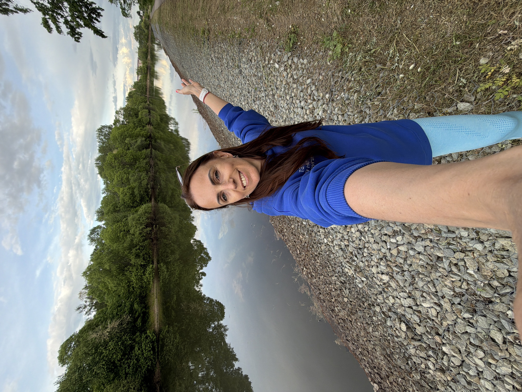 A woman takes a selfie outdoors next to a body of water in Birobidzhan, Russia. She has long brown hair, is smiling cheerfully at the camera, and wears a royal blue long-sleeved top and light blue patterned leggings. Sunglasses are perched on her head, and a white smartwatch is visible on her left wrist. Her right arm is extended behind her, gesturing towards the scenic background.
The setting features a calm body of water, likely a river or canal, stretching into the distance. The bank where she stands is composed of grey gravel or rocks secured by a wire mesh, with some patches of grass. On the opposite side of the water, a dense line of lush green trees forms a natural backdrop, with their reflections visible in the water. The sky is partly cloudy, suggesting the soft light of late afternoon or early evening. Cyrillic text, "та не я не друга," is visible on her blue top. The overall scene captures a moment of happiness in a serene natural environment.