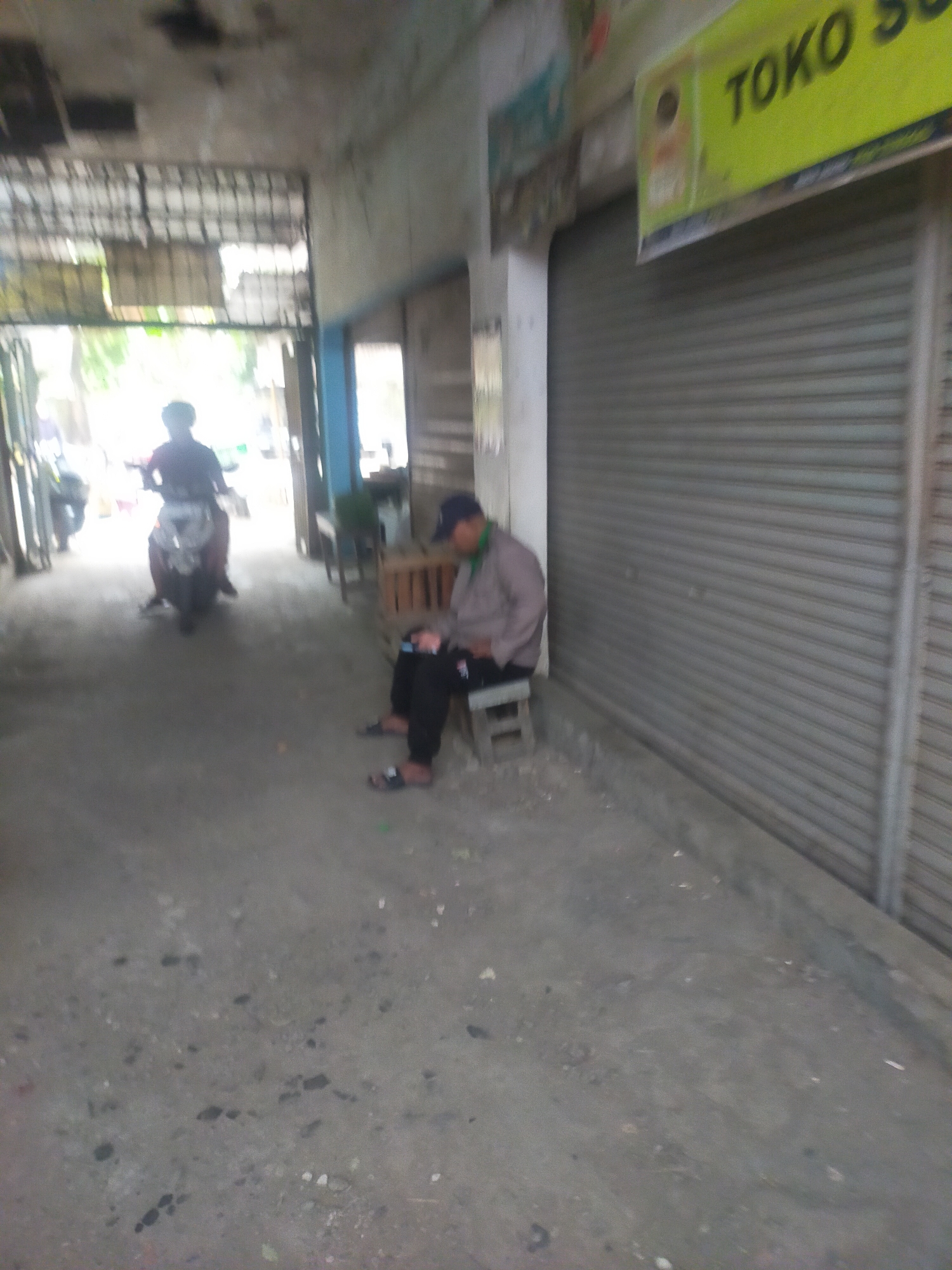 The image shows a street scene in Trayeman, Indonesia. A person is sitting on a small stool on the right side of the image, looking at their phone. They are wearing dark pants, a light-colored long-sleeved shirt, and a dark baseball cap. In the background, a person is riding a motorcycle. The street is paved and appears to be in a commercial area, with storefronts on the right side of the image, one of which has a sign that reads "TOKO SU." The environment is somewhat dilapidated, with debris on the ground.