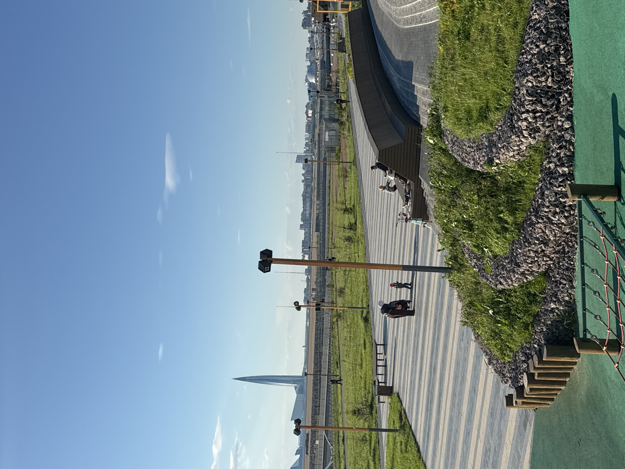 The image depicts a park or public space in Saint Petersburg, Russia, featuring a pedestrian walkway with alternating light and dark gray paving stones. Along the walkway are modern streetlights with rectangular fixtures. In the background, the Lakhta Center, a prominent skyscraper, is visible. People are present on the walkway, including individuals walking and sitting on benches. Adjacent to the walkway is a landscaped area with grass, plants, and a retaining wall constructed of rocks in wire mesh. The sky is clear and blue. To the right of the walkway is an artificial slope with vegetation and a playground structure with wooden steps and netting.