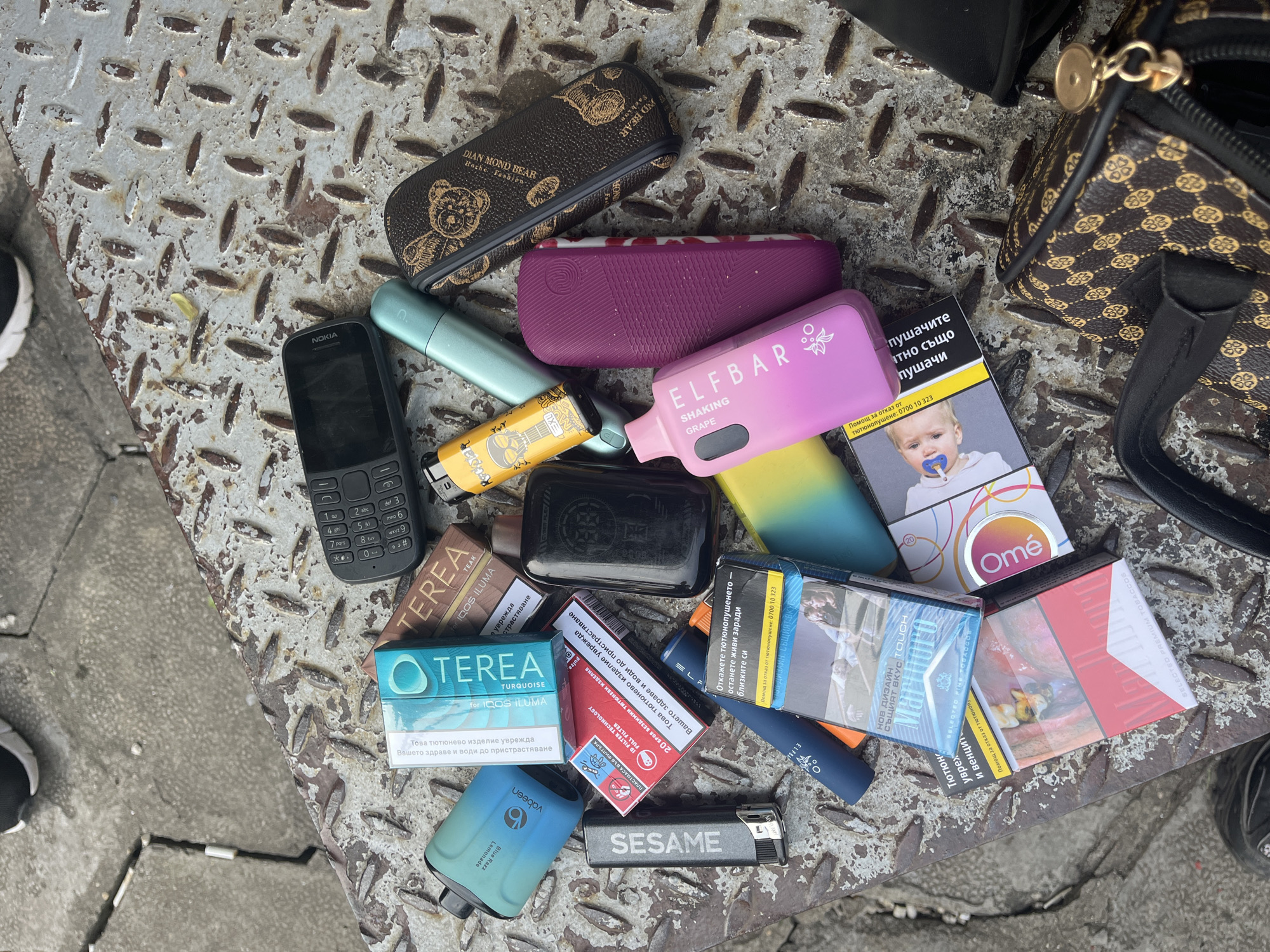A collection of personal items, predominantly nicotine products, lies scattered on a heavily worn, metallic diamond-plate surface, likely an urban ground fixture. The setting appears to be outdoors in Sofia, Bulgaria, given the Bulgarian text on many products.
The array of objects includes a black Nokia feature phone, several vaping devices (a pink "ELFBAR SHAKING GRAPE," a blue "vabeen Blue Razz Lemonade," and other unidentified vapes), and various tobacco products. There are packs of heated tobacco sticks like "TEREA TEAK for IQOS ILUMA" and "TEREA TURQUOISE for IQOS ILUMA." Numerous cigarette packs are visible, some featuring graphic health warnings common in Bulgaria, such as a crying baby with a pacifier and decaying teeth, alongside texts like "Откажете тютюнопушенето - останете живи заради близките си" (Quit smoking - stay alive for your loved ones) and "Помощ за отказ от тютюнопушене: 0700 10 323" (Help to quit smoking: 0700 10 323). Other visible brands include "Omé" and a red pack labeled "SELECTED FINE TOBACCOS."
Also present are two lighters (one black with "SESAME," another yellow with a graphic of a guitar player), two glasses cases (one brown with a "DIAN MOND BEAR Hache Fashion" logo and one plain purple), and part of a small brown patterned handbag. The items seem to be casually placed or possibly momentarily set down on the rough, textured metal surface during daytime. The diversity of products, from traditional cigarettes to heated tobacco and vapes, reflects various contemporary nicotine consumption habits.