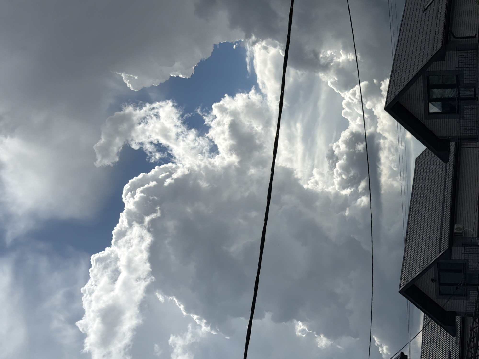 The image depicts a partly cloudy sky with a building on the right side. The sky features a mix of light and dark clouds, with a patch of blue sky visible in the upper left. Two power lines are stretched across the center of the image, partially obscuring the clouds. The building on the right has a multi-tiered, angular design with textured, dark grey cladding. The specific location in Rossiyskiy, Russia is not discernible from the image.