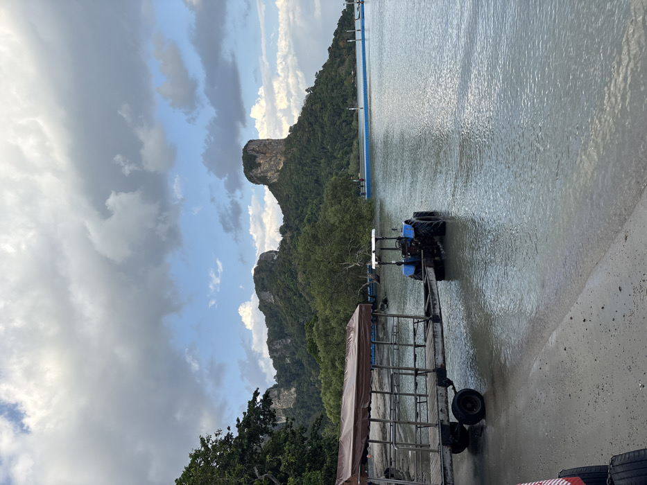 The image depicts a coastal scene in Ban Ao Nang, Thailand, under a sky with significant cloud cover and patches of blue. In the foreground, a sandy beach transitions into calm, light green-blue water. A large, four-wheeled trailer with a brown canvas canopy and metal frame rests on the wet sand and extends partially into the water, positioned to the left. Immediately behind the trailer and partially submerged in the water is a blue tractor, facing away from the camera, with its rear wheels fully submerged. In the midground, the calm water extends towards a distant shoreline densely covered with green trees and mangroves. A blue floating dock or walkway, supported by white vertical poles, extends into the water from the right side of the frame, with indistinct human figures visible near its base. The background is dominated by large, steep limestone karst formations (mountains/cliffs) covered in lush green vegetation, characteristic of the region's topography. The scene suggests an operation involving the tractor and trailer for moving objects or people to or from the water.
