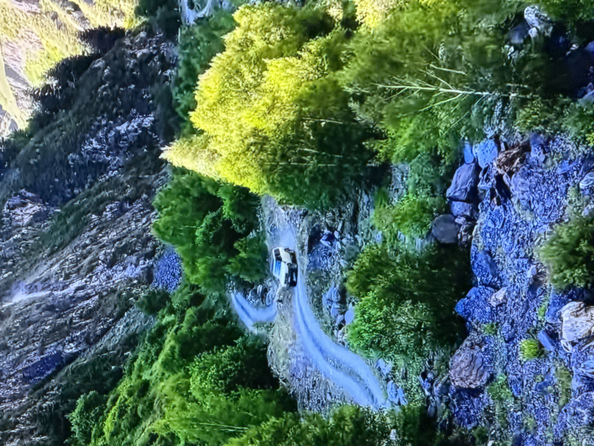 A white SUV drives on a winding dirt road in a mountainous, green landscape. The road curves around a steep, rocky mountainside covered in dense green foliage and trees. The mountains in the background are steep and rocky, with patches of greenery. The sunlight illuminates some of the trees, giving them a bright yellow-green hue. The overall scene depicts a remote, natural environment.