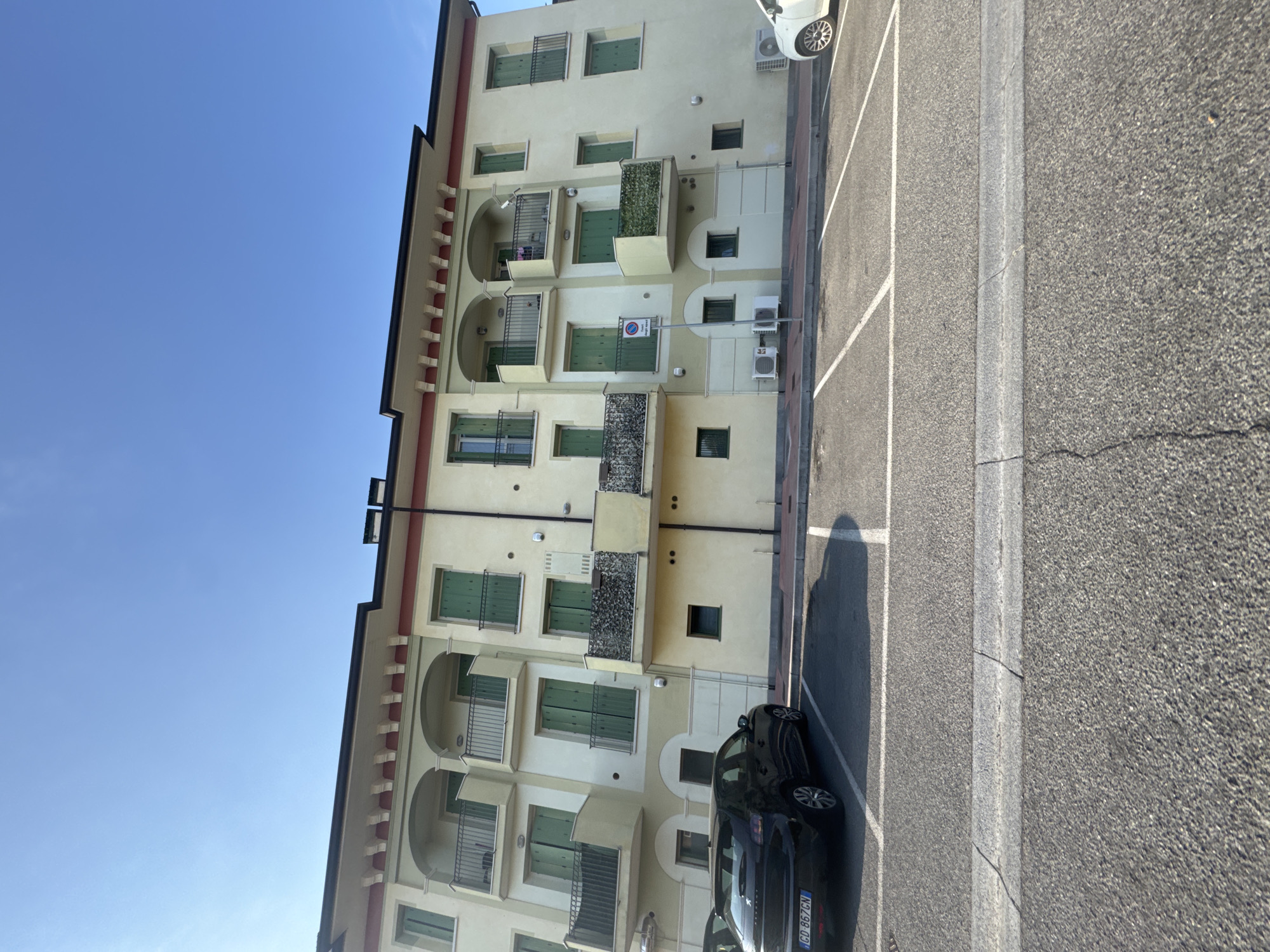 A low-angle, eye-level medium shot captures the exterior of a multi-story building with a pale yellow facade and red trim, under a clear blue sky. The building features several balconies with green shutters on its windows. In the foreground, a black hatchback car is parked in a lot marked with white lines, with its rear end closest to the camera. To the right, a white car is partially visible. The parking lot is paved with asphalt, and a concrete curb separates it from a sidewalk. A blue and white traffic sign, possibly indicating parking regulations, is visible on a pole in front of the building. The building appears to be an apartment complex.