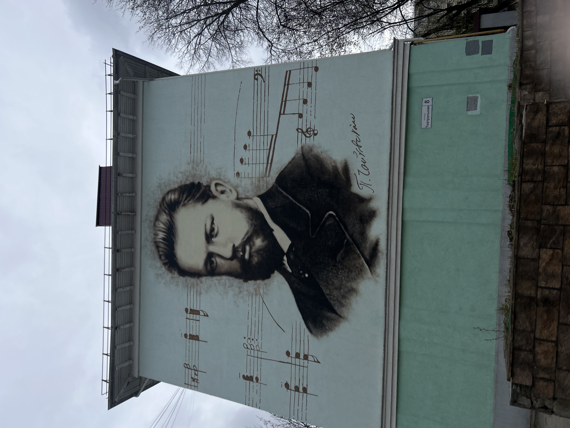 The image displays a large mural painted on the side of a building in what appears to be an urban setting, consistent with Klin, Russia. The central subject of the mural is a sepia-toned or dark brown portrait of a bearded man, identifiable as the renowned composer Pyotr Ilyich Tchaikovsky. His signature, "П. Чайковский" (P. Tchaikovsky), is prominently displayed in cursive script to the right of his head. He is depicted with a contemplative or somewhat somber expression, looking slightly to his left.
The background of the portrait features multiple musical staff lines with various notes, G-clefs, and a bass clef, emphasizing his musical legacy. The mural is painted on a light-colored, possibly off-white, section of the building.
Adjacent to the mural on the right is another section of the building painted in a pale green color. On this green wall, a rectangular street sign is visible, bearing the text "Театральная" (Teatral'naya), which translates to "Theatrical Street," along with the house number "8".
Above the building, bare tree branches are visible against an overcast, light gray sky, suggesting either winter or early spring conditions. The top of the building features a simple pitched roofline. To the far right, part of another building with a stone or brick facade can be seen. The image captures a blend of public art, architectural elements, and natural surroundings under a muted sky.