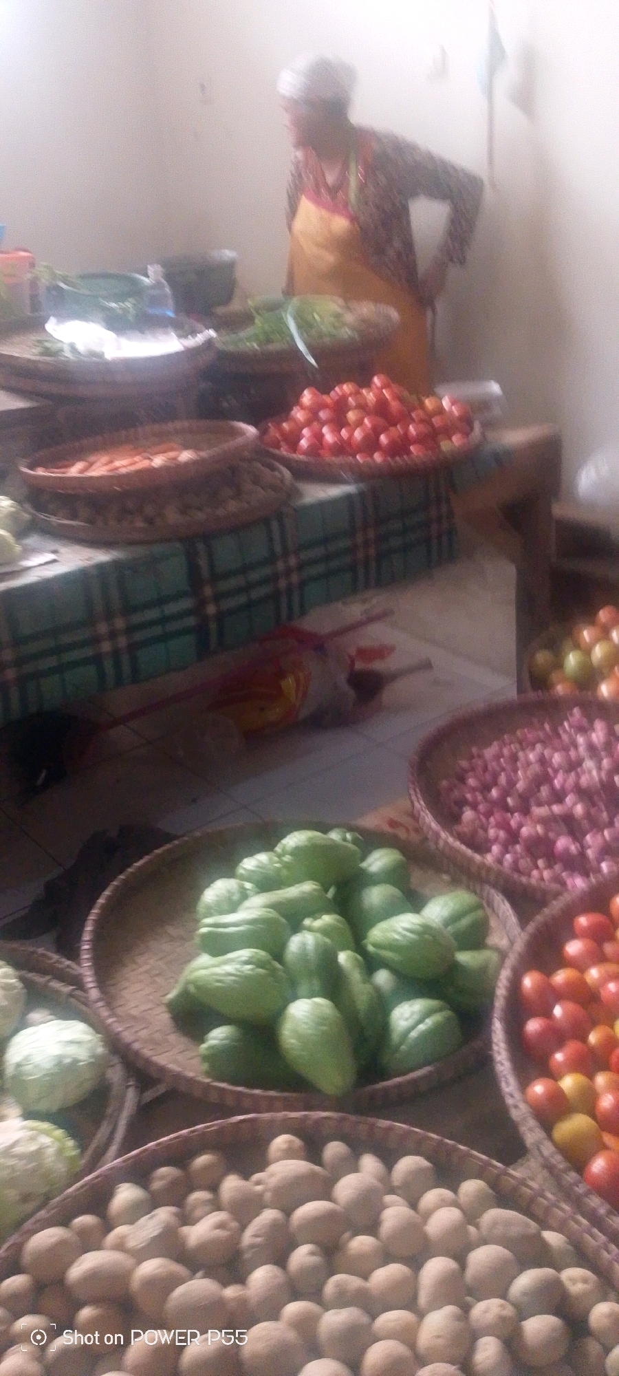 The image shows a market stall with a variety of fresh produce. In the foreground, there are baskets filled with potatoes and green chuchus. To the right, a basket overflows with red tomatoes, and another contains numerous small red onions. In the background, a woman wearing an apron stands behind the produce. The stall is set up on a table covered with a green and white checkered cloth. The environment appears to be an indoor market.