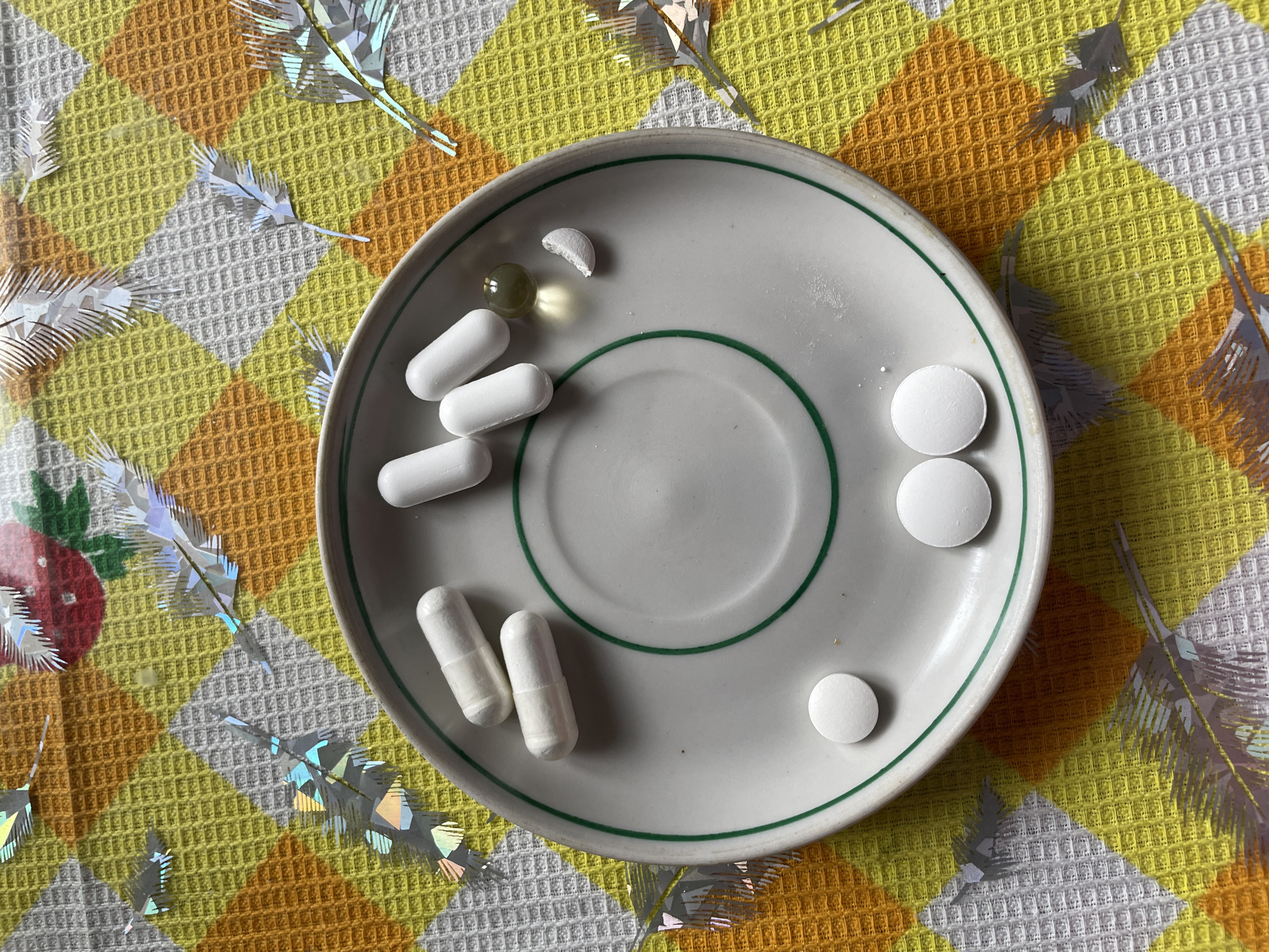 The image shows a small, white plate with a green rim, sitting on a yellow, orange, and white checkered tablecloth. On the plate are several pills: three long, white capsules; two round, white pills; one single, round, white pill; one yellowish, spherical capsule; and a small piece of a broken white pill. Decorative, shiny, feather-like objects are scattered around the plate on the tablecloth. The image does not provide information to determine the location is in Talmenka, Russia or to provide other details about people, subjects, actions or interactions.