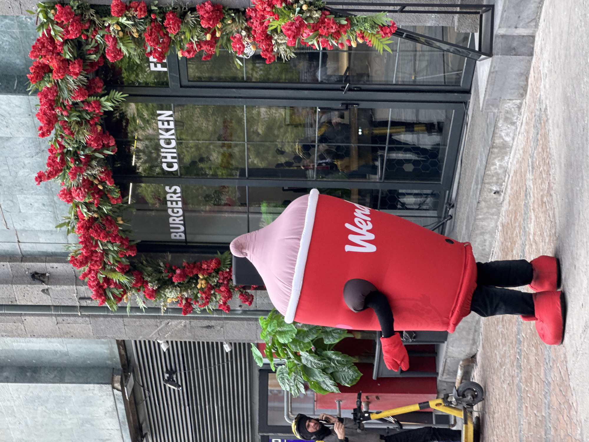 An outdoor, daytime shot captures a person dressed in a red and pink Wendy's "Frosty" cup mascot costume standing on a sidewalk in front of a Wendy's restaurant in Yerevan, Armenia. The mascot, prominently displaying the "Wendy's" logo in white script on its red body, features a light pink, swirled top resembling whipped cream, black arm sleeves, red gloves, black pants, and large red boot-like shoes.
Behind the mascot is the entrance of a building, which features modern glass doors and windows. A vibrant, thick garland of red flowers and green foliage frames the entrance, adding a festive touch. Visible white text on the glass windows reads "BURGERS" and "CHICKEN", with a partial "F" (likely for "Fries" or "Frosty") also visible, confirming the establishment's offerings. The building exterior appears to be constructed of gray stone or concrete.
On the left side of the frame, near the mascot's feet, a yellow electric scooter is parked on the paved ground. Partially visible behind the scooter is another person, wearing dark clothing and a black head covering, looking downwards. The scene suggests a promotional event or a typical day outside the fast-food establishment in an urban setting.
